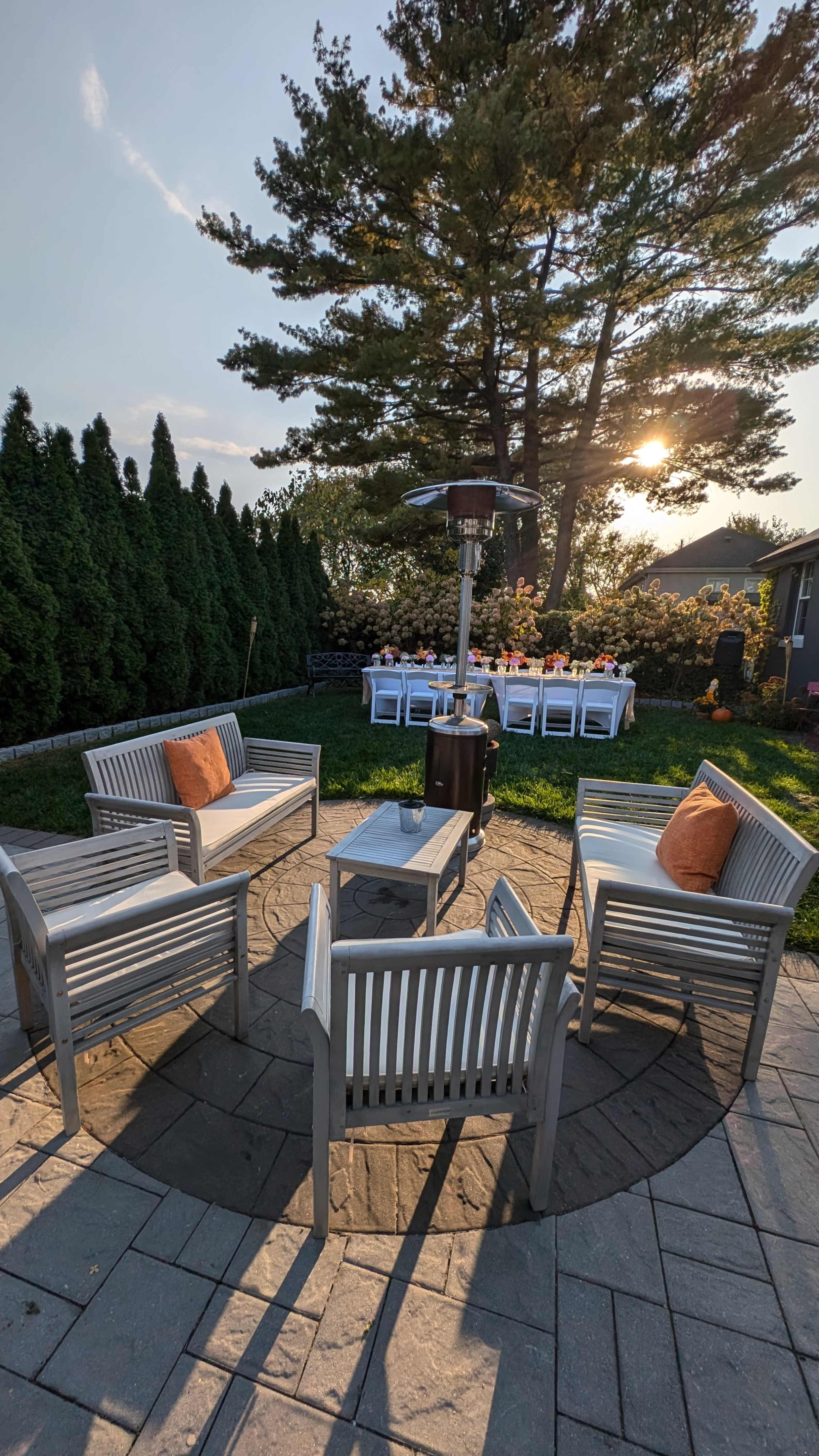 A circular seating area with four chairs and a table is set up around a patio heater in a backyard, with a dining table in the background and a setting sun behind trees.