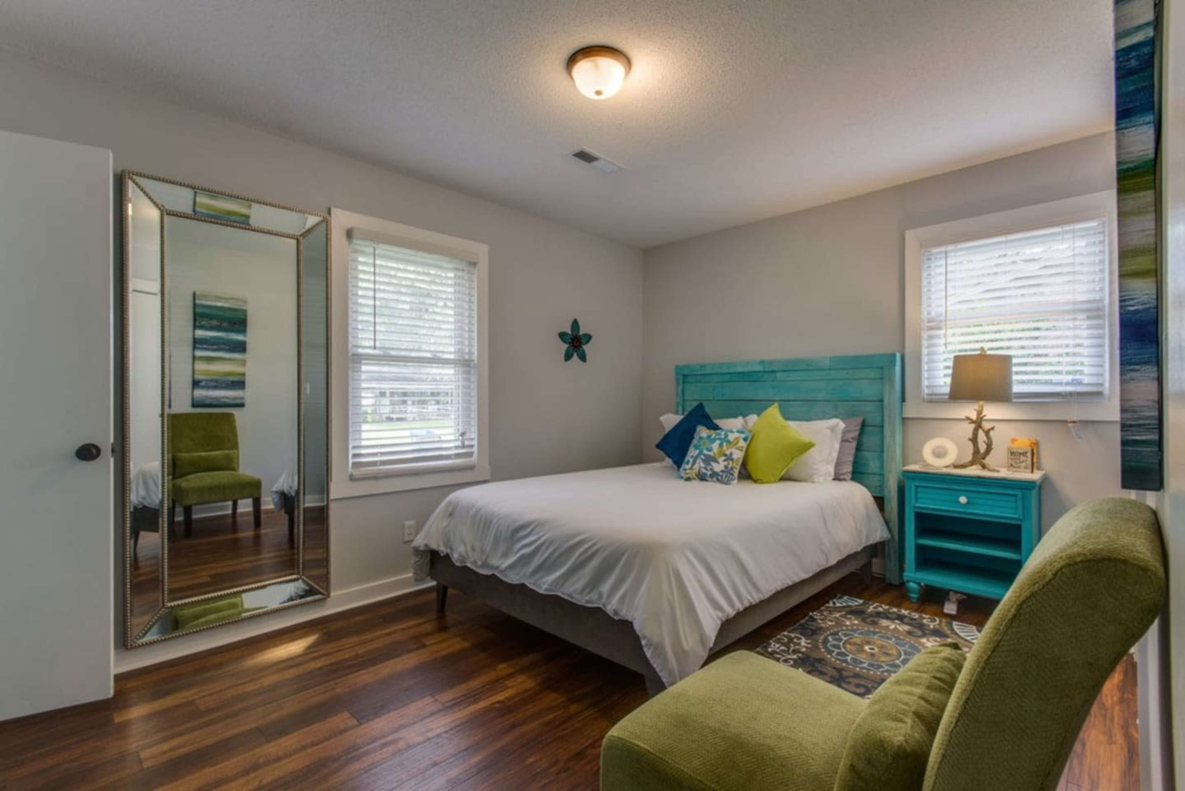 A cozy bedroom featuring a bed with colorful pillows, a green chair, a mirror, and a small bedside table.