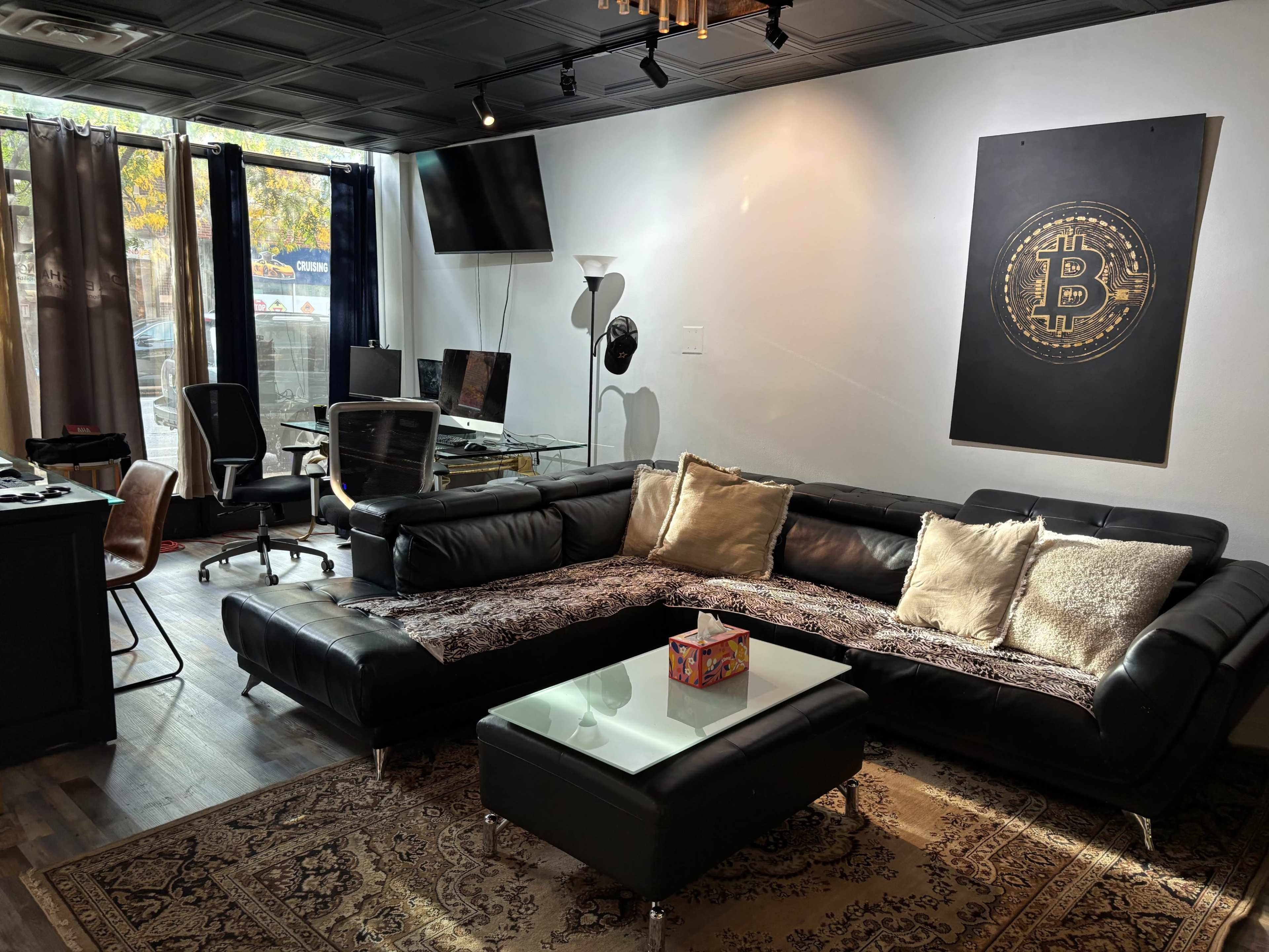 The image shows a modern office lounge area with a black sectional sofa, a coffee table, a large wall art featuring a Bitcoin symbol, and workstations equipped with computers.
