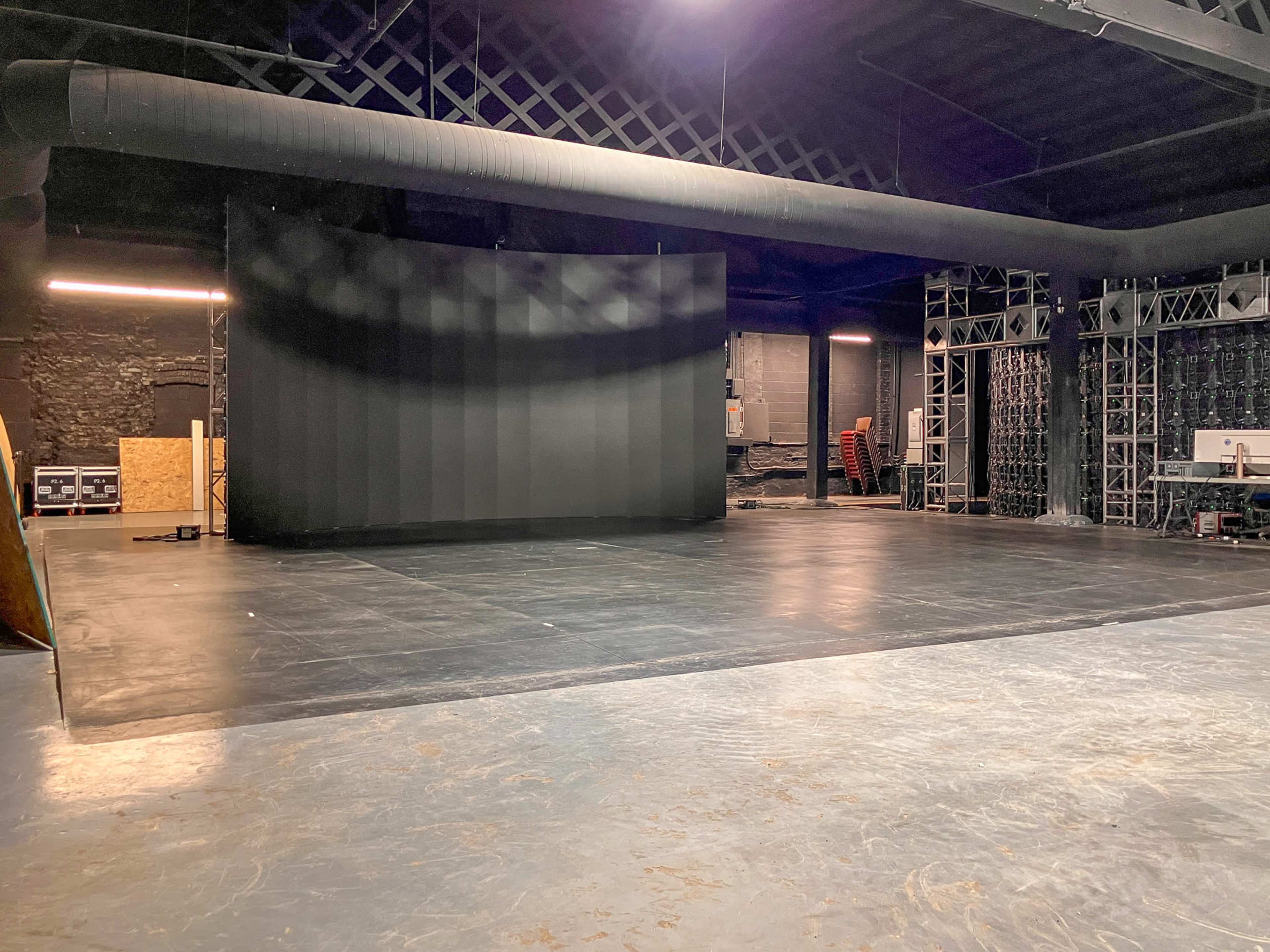 The image shows an empty stage area with a large black backdrop and industrial equipment arranged along the walls in a dimly lit space.