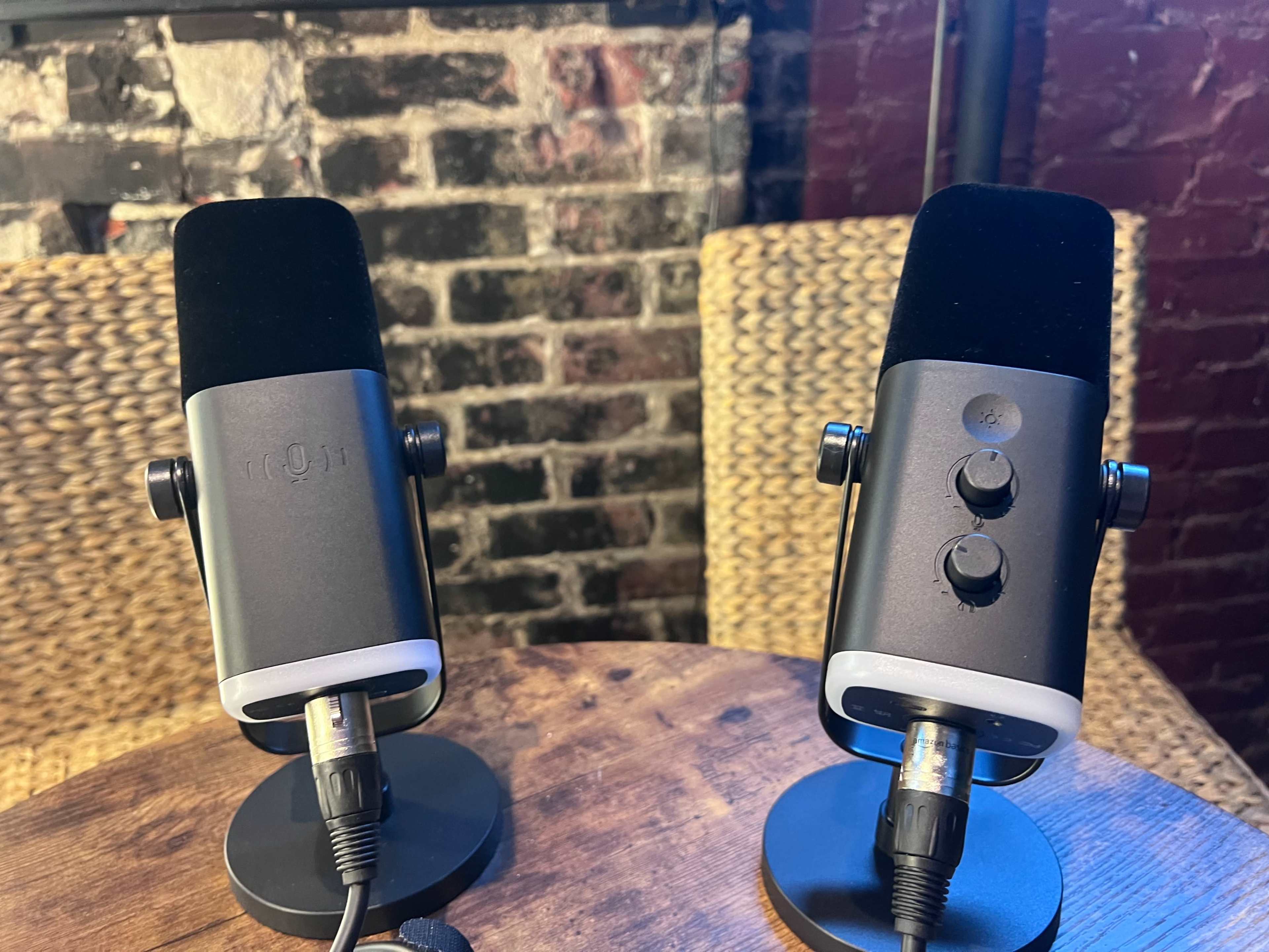 Two microphones are placed on a wooden table with a brick wall background.