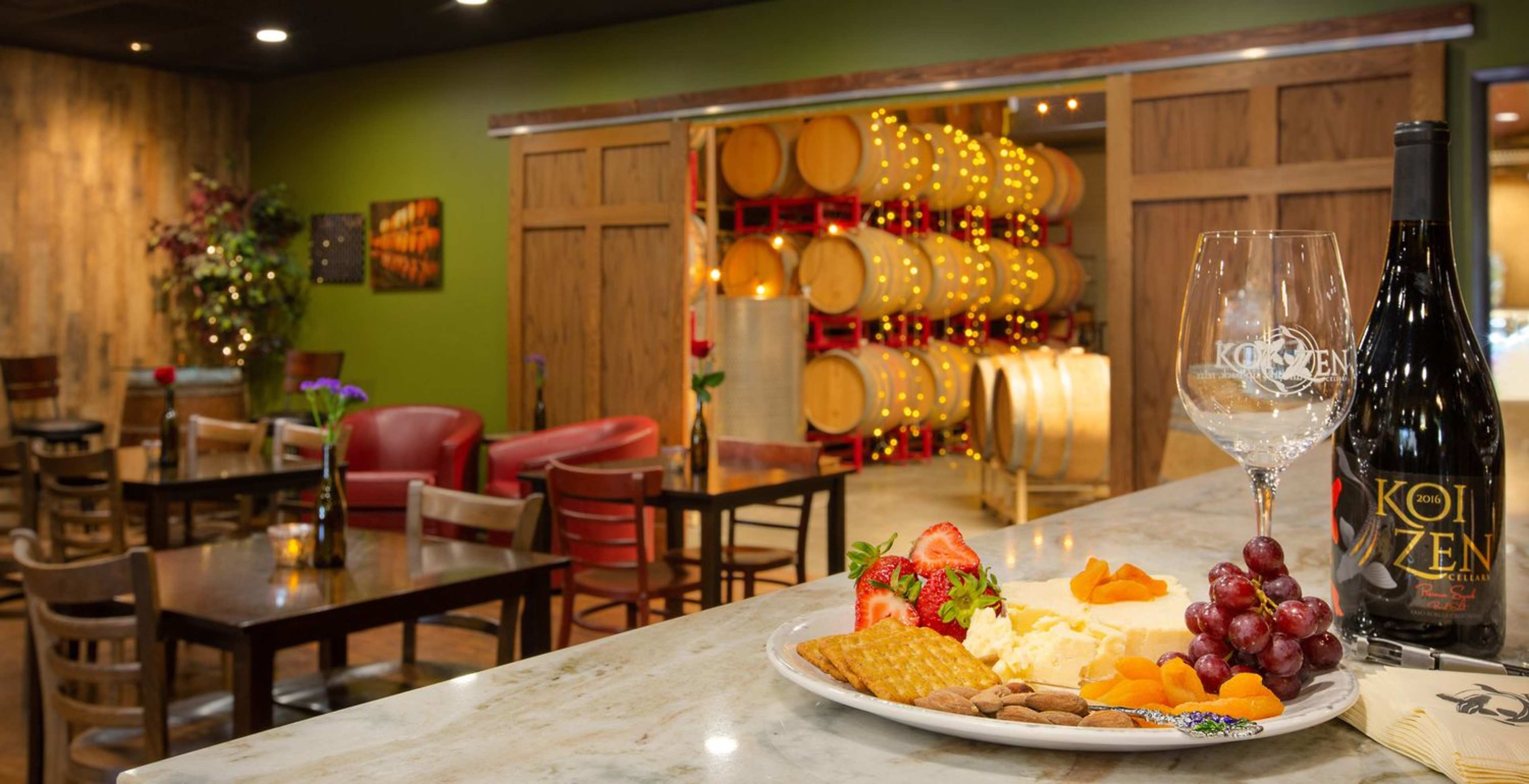 A wine tasting room with a plate of cheese, fruits, and nuts in the foreground, and wooden wine barrels in the background.