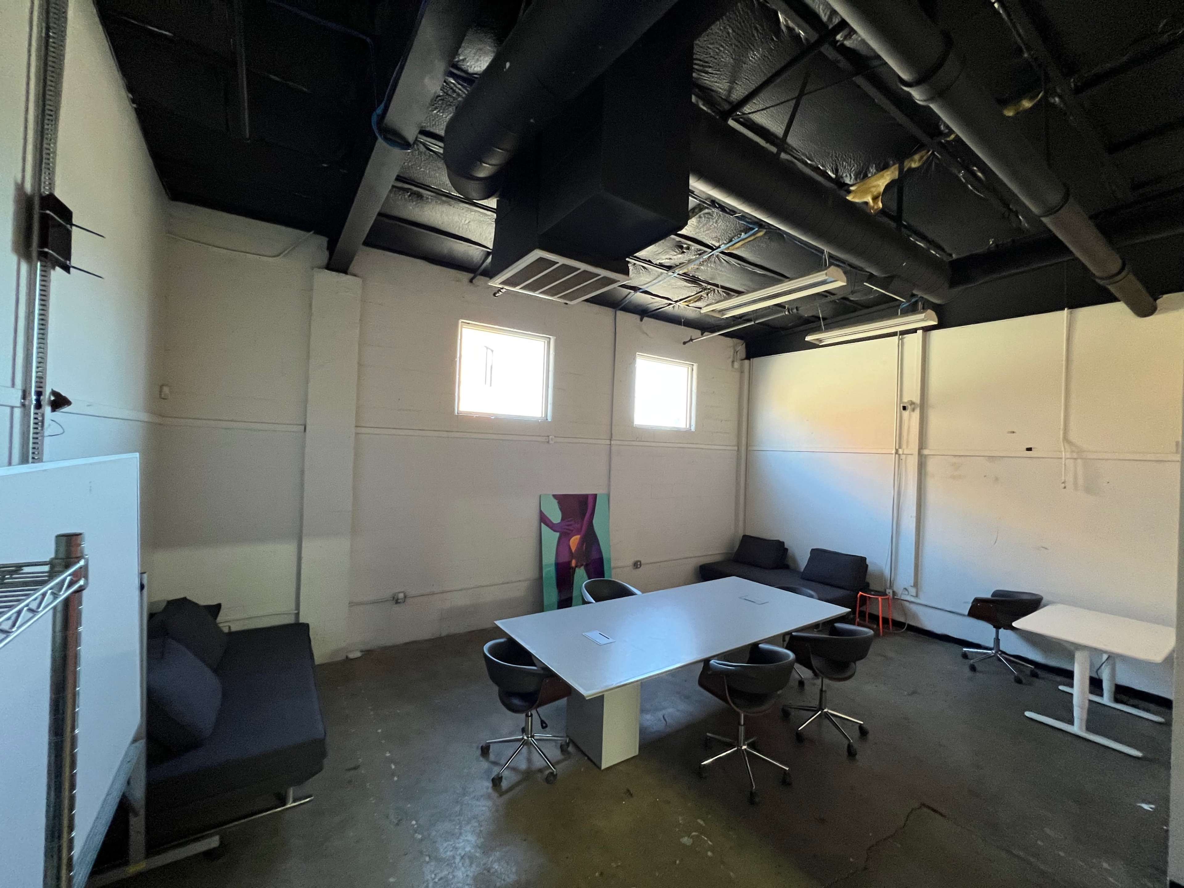 The image shows a sparsely furnished office space with a large table surrounded by chairs, a few couches, and two windows providing natural light.