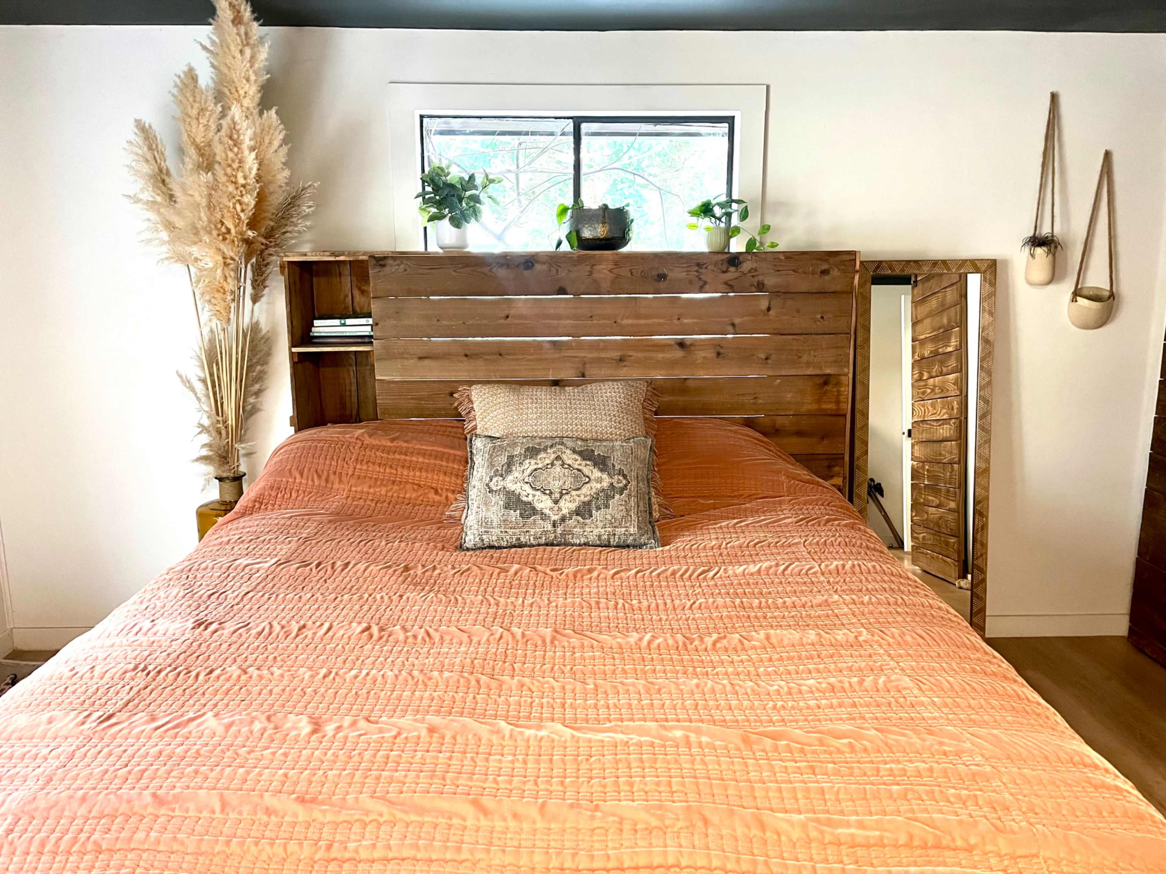 The image shows a cozy bedroom featuring a wooden bed frame with a rust-colored quilt, a decorative pillow, and plants on a shelf behind the headboard.