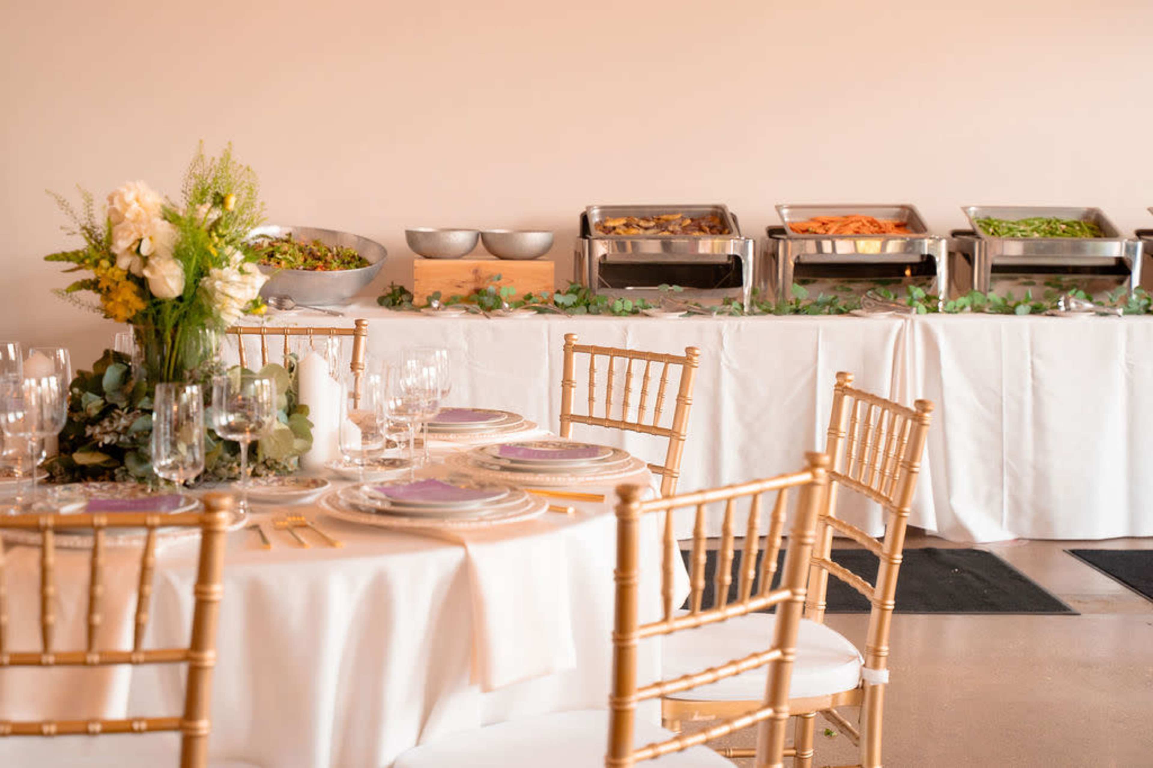 A dining setup with golden chairs, elegantly arranged tables, and a buffet featuring a variety of dishes in chafing dishes.