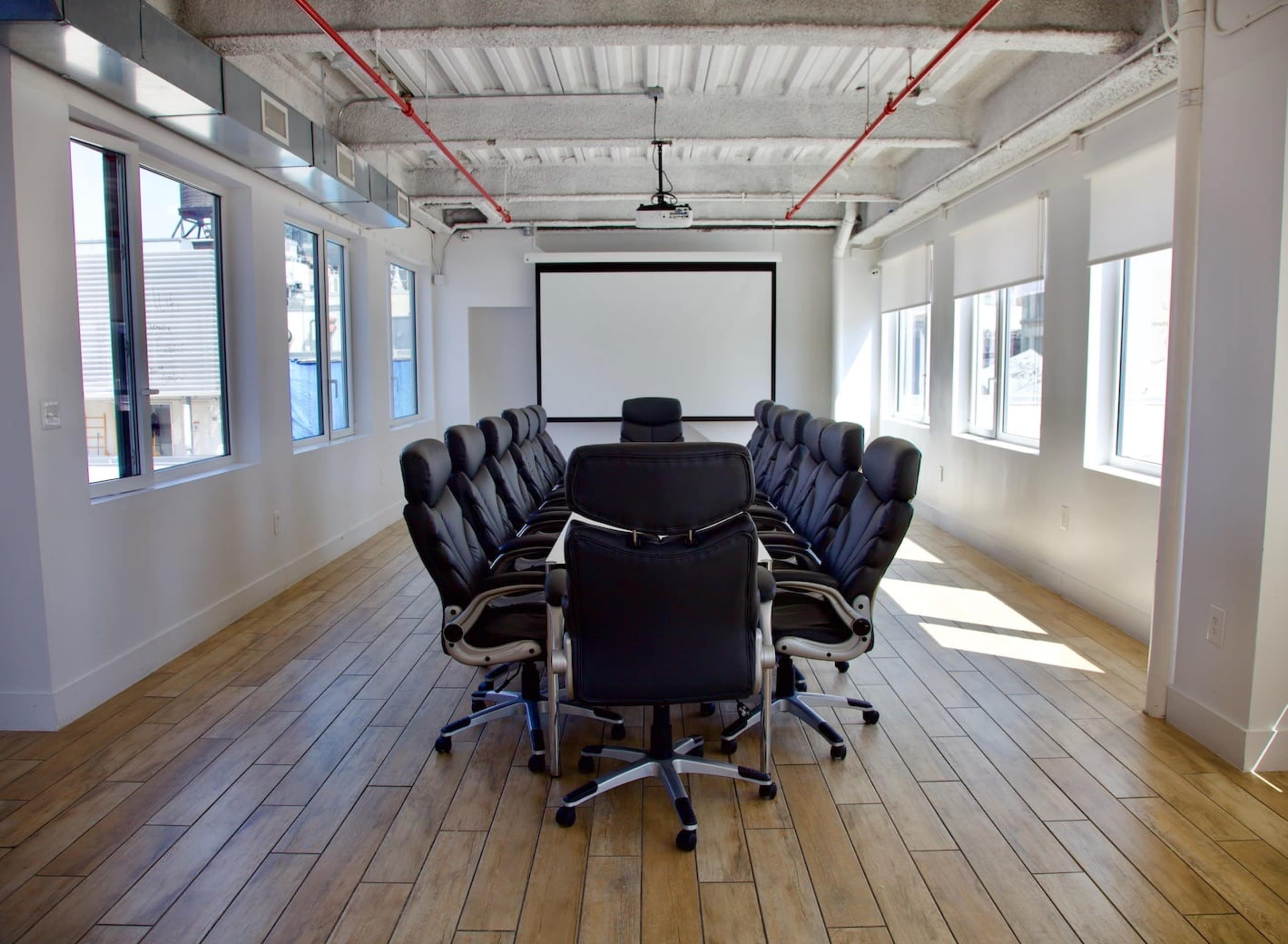 A modern conference room features a long table surrounded by black ergonomic chairs, with large windows and a projector screen at one end.