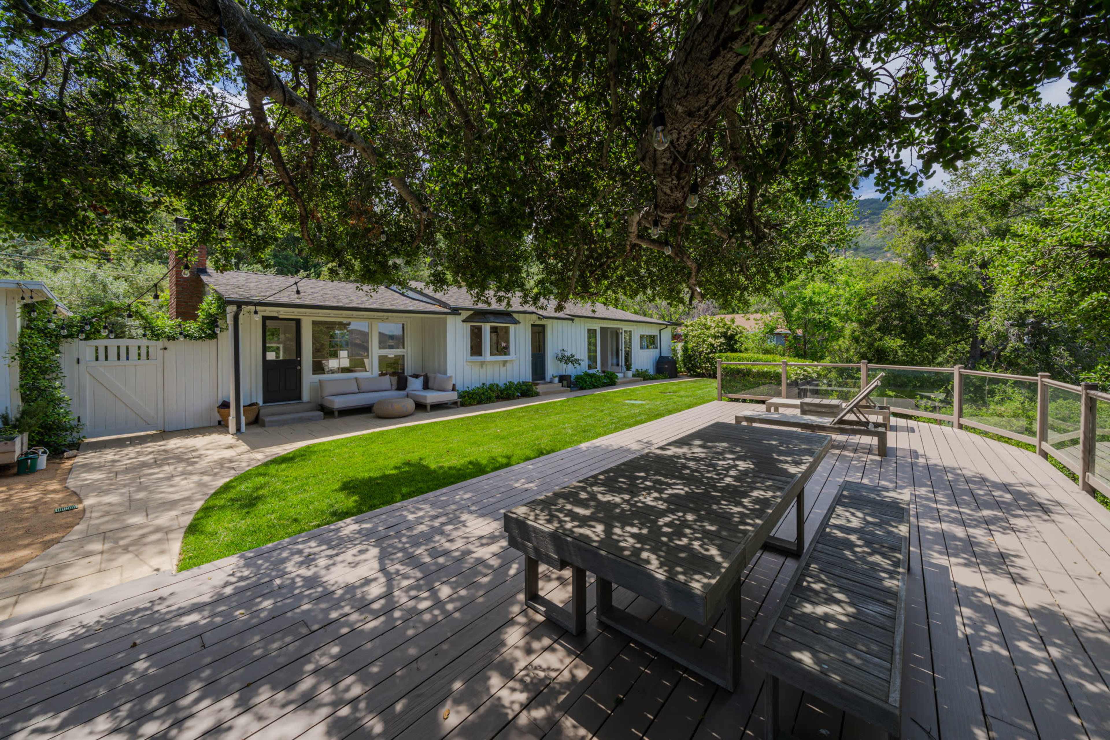 The image shows a modern house with a large wooden deck and a spacious lawn surrounded by trees.