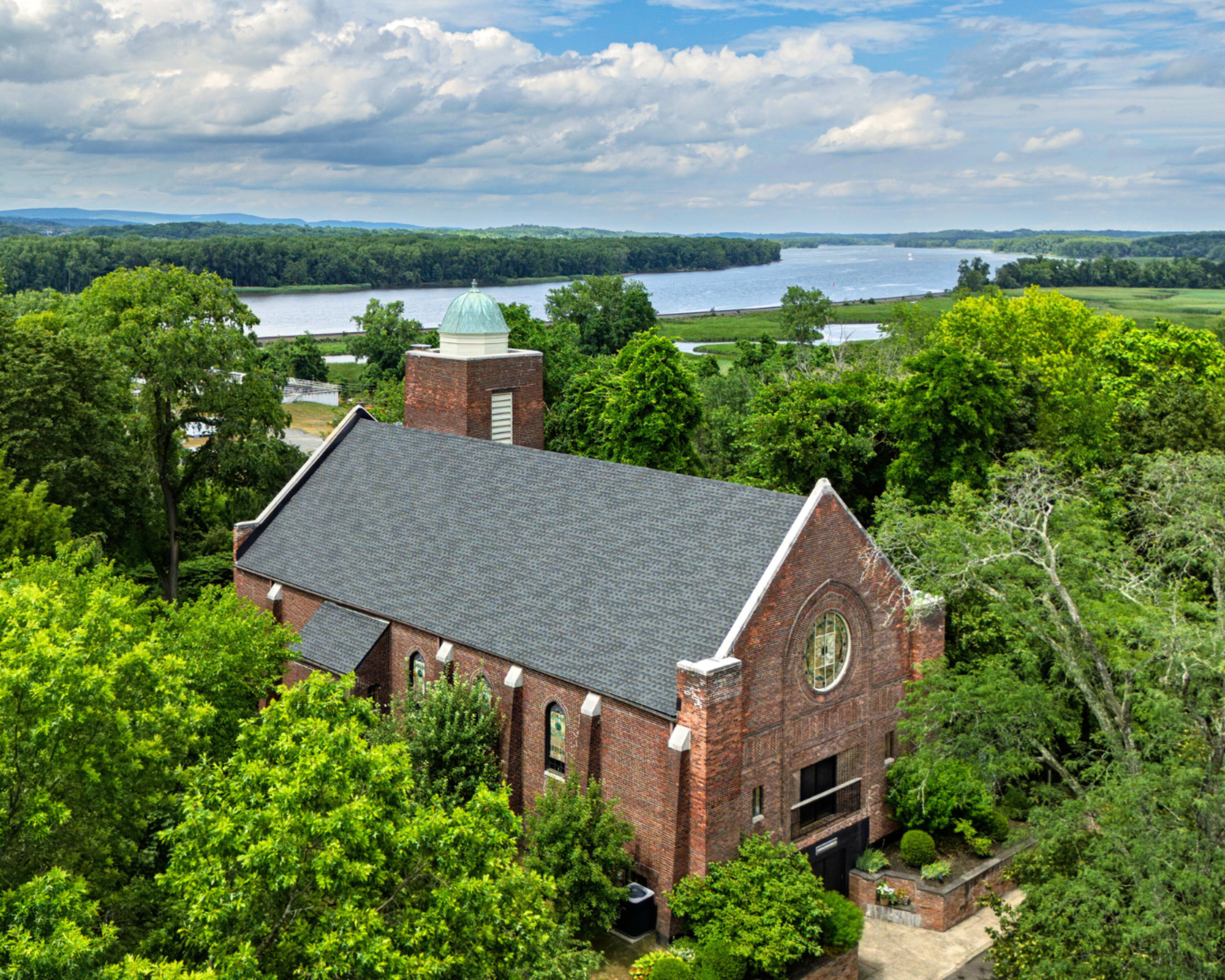 A brick church with a dome is surrounded by lush greenery and overlooks a river.