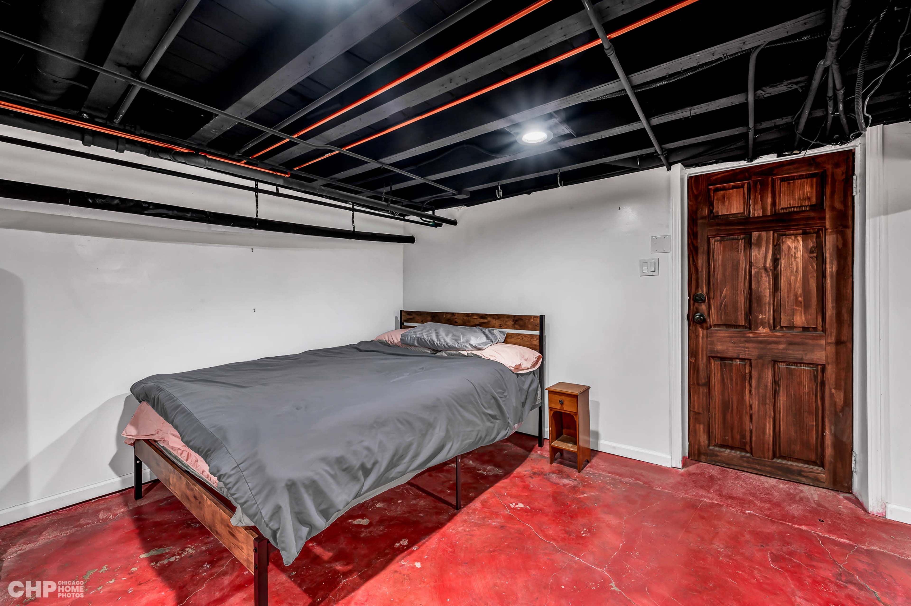 A basement bedroom with a wooden bed, gray bedding, a small nightstand, and a wooden door, all set against a white wall and red concrete floor.