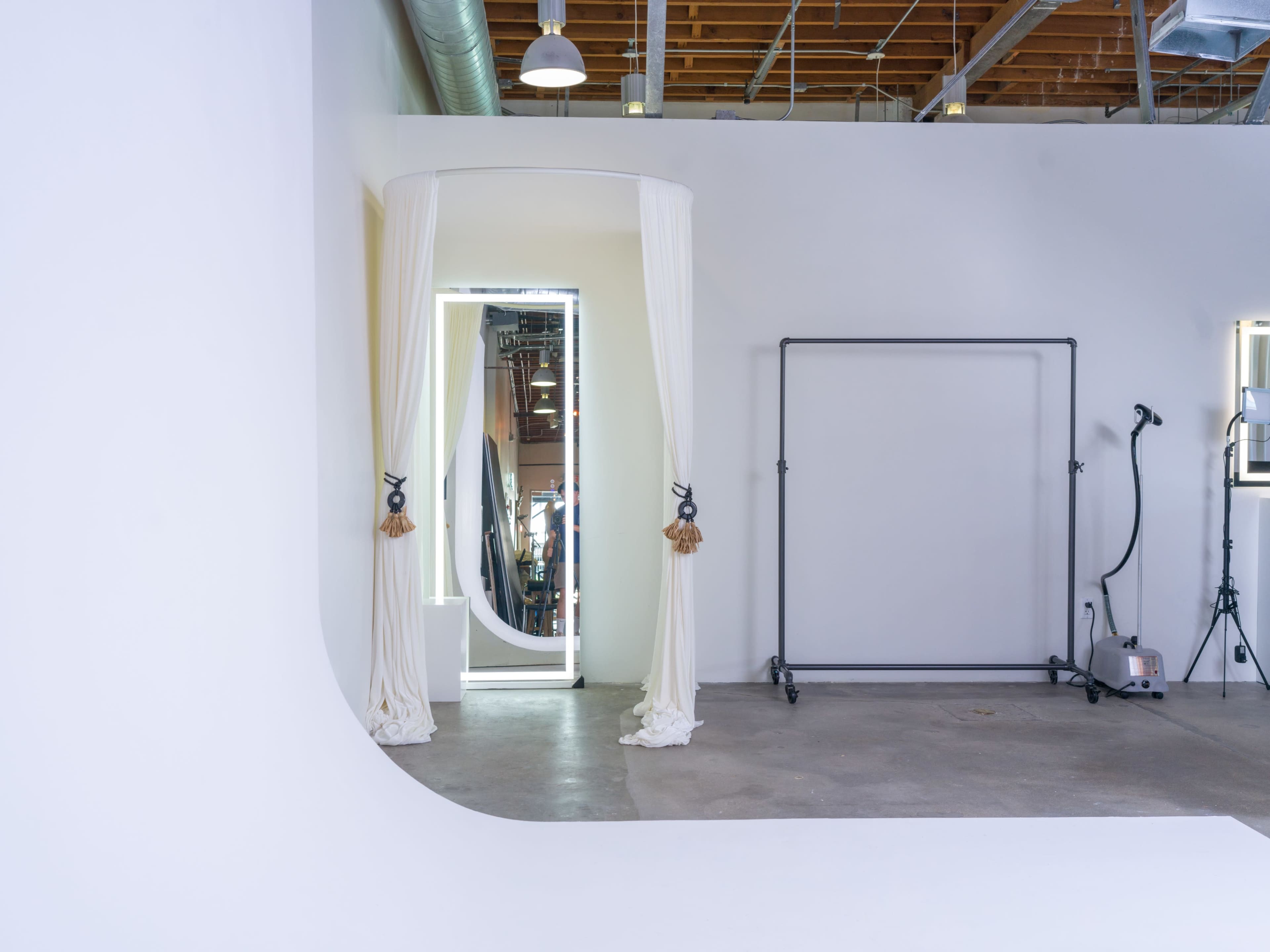 A minimalist studio space with a curved white backdrop, an illuminated mirror framed by curtains, and a metal structure against the wall.