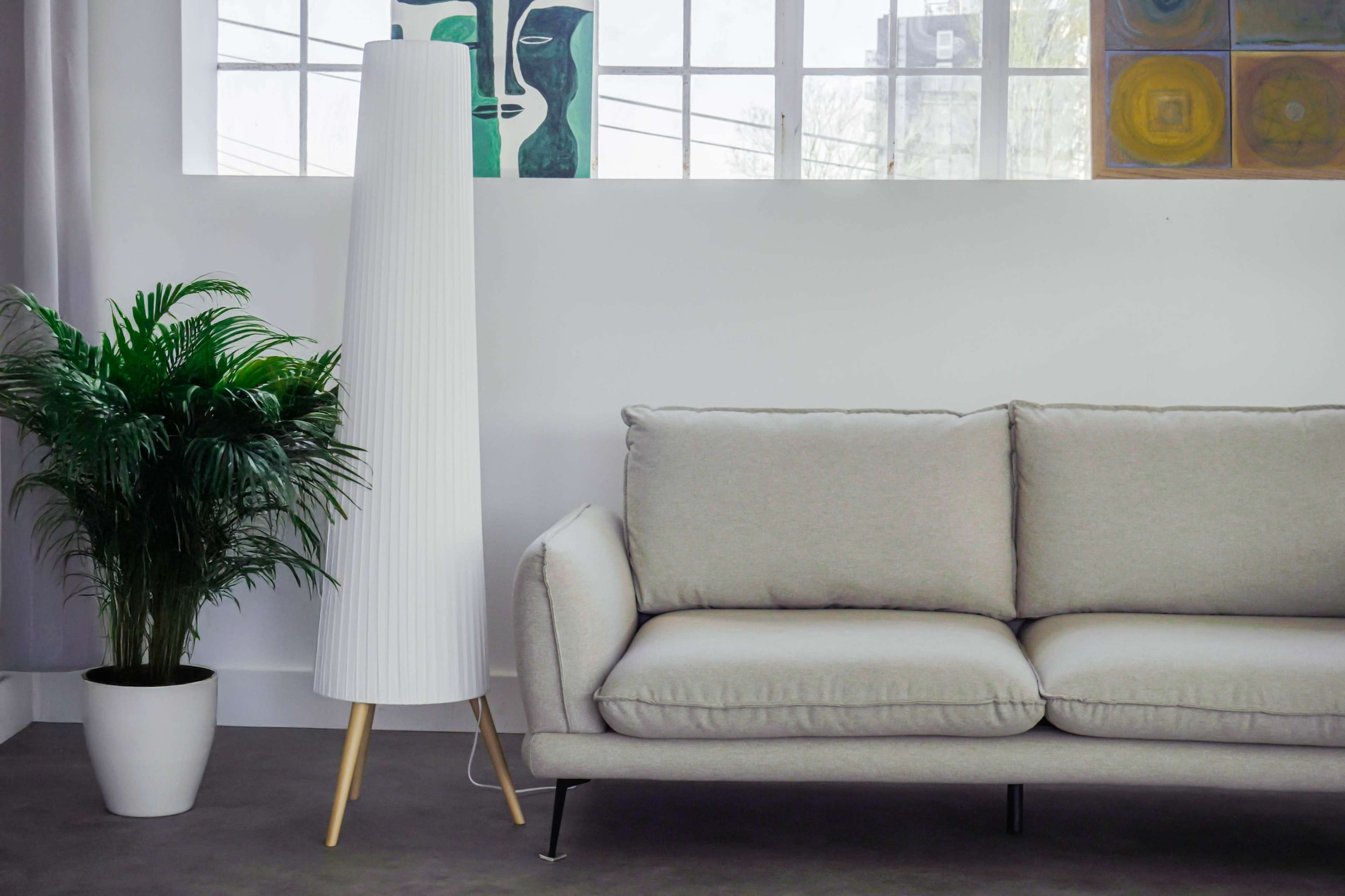 The image shows a modern living room setup featuring a light gray sofa, a tall white lamp, a potted plant, and artwork on the wall.