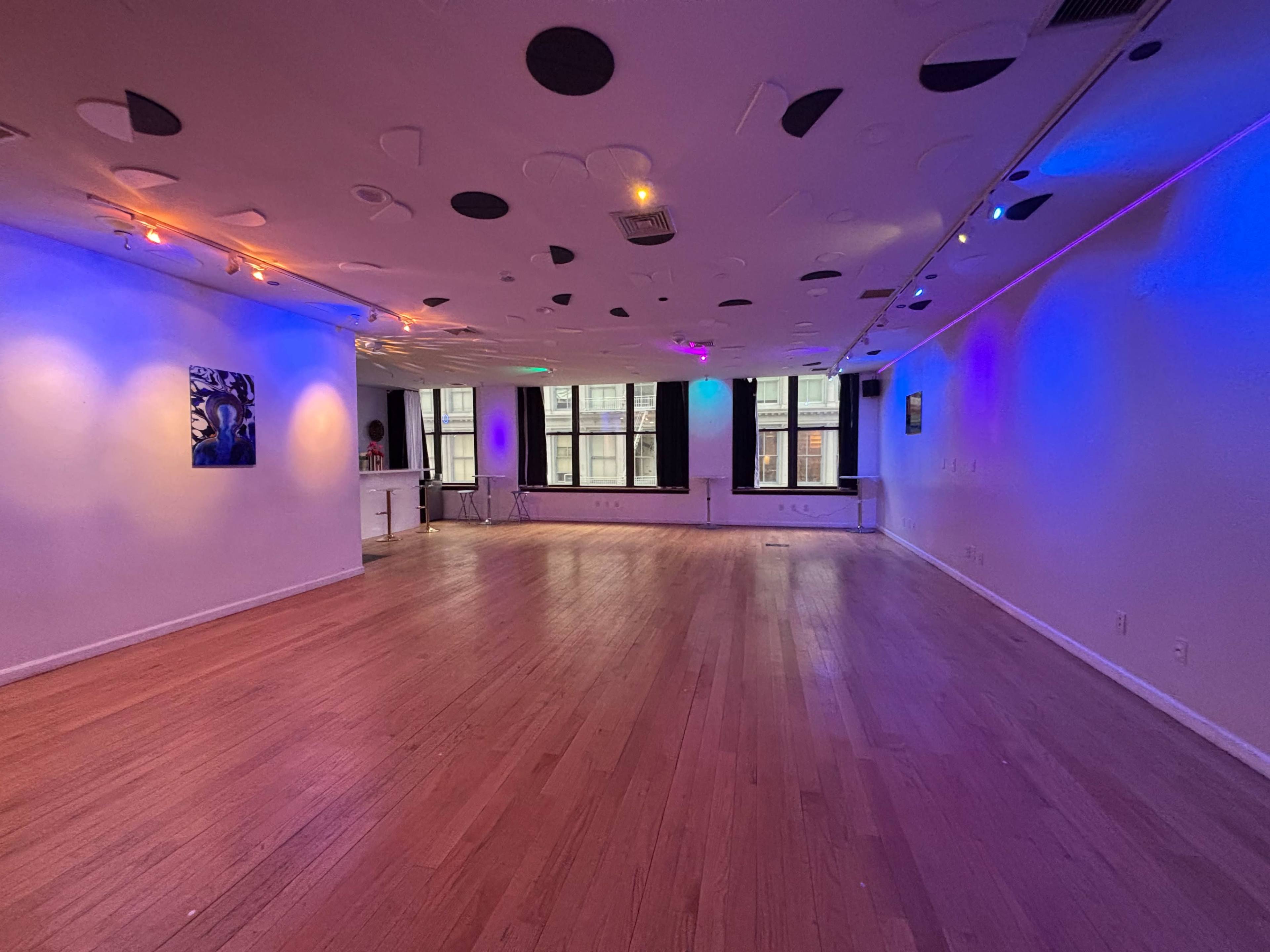 A spacious, well-lit room features wooden flooring and colorful lighting with abstract wall art.