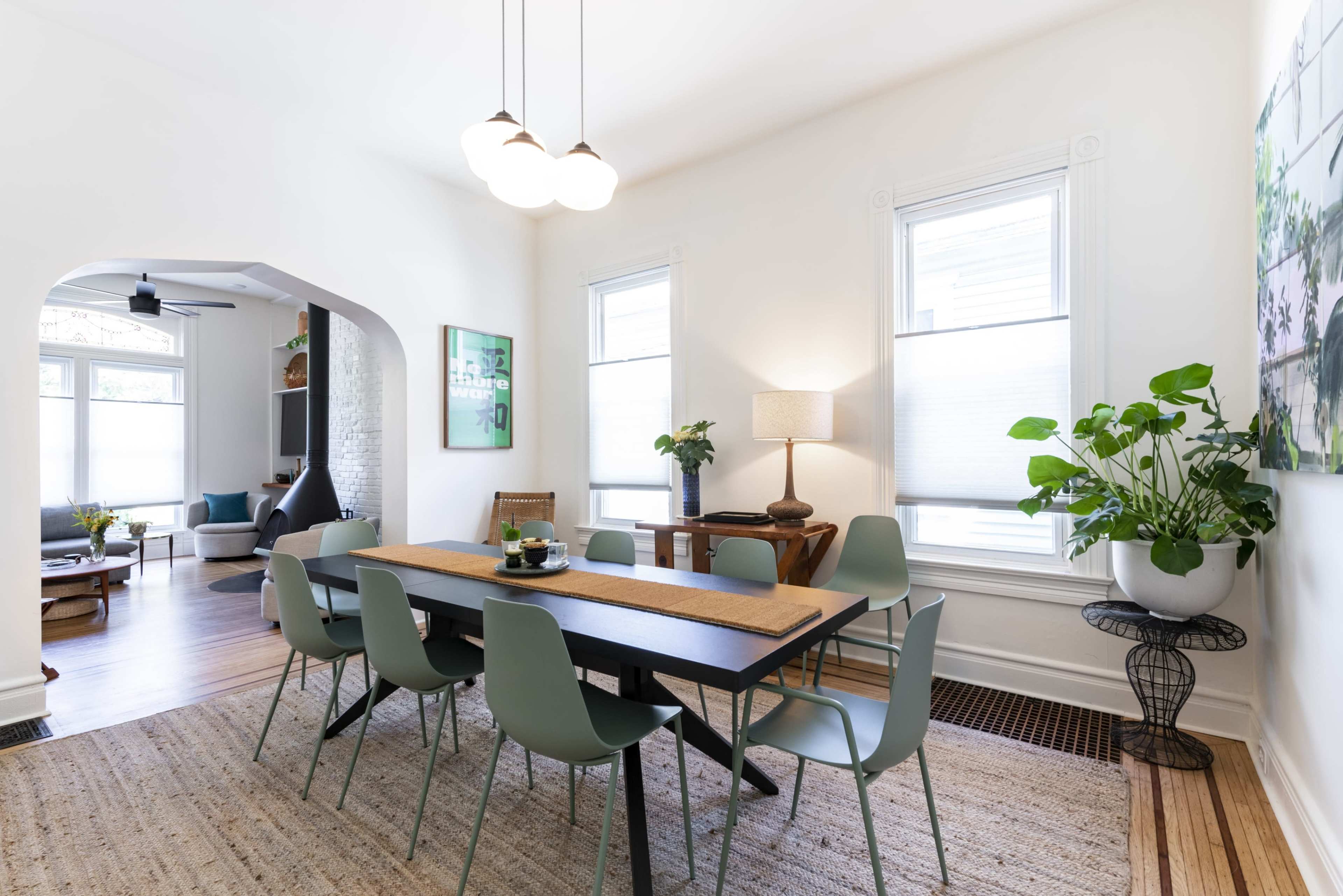 A spacious dining area features a long table surrounded by modern chairs, with large windows letting in natural light and plants adding greenery to the scene.