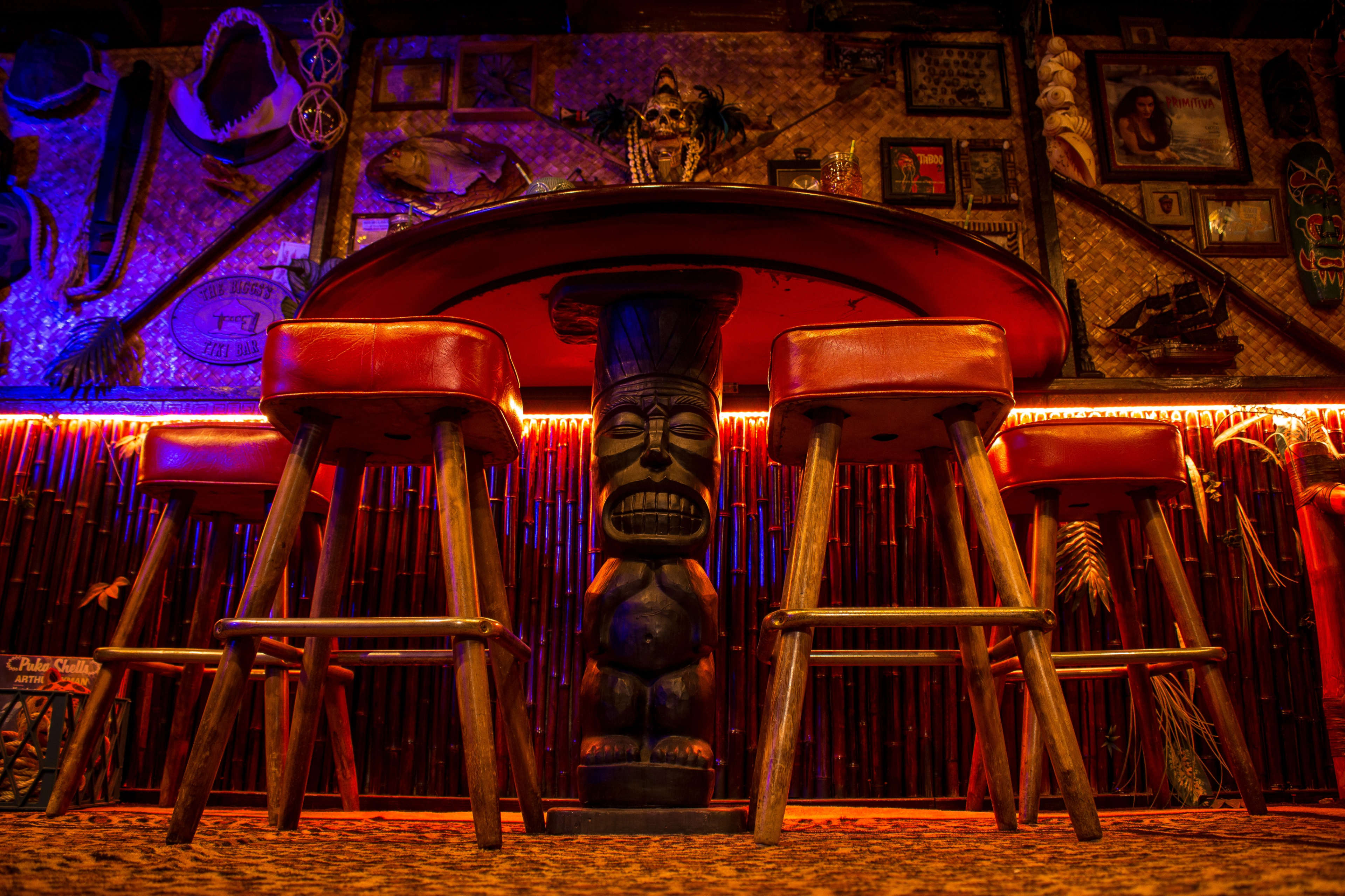 A tiki bar scene features a round red table surrounded by wooden stools, highlighted by colorful ambient lighting and tropical decor on the walls.