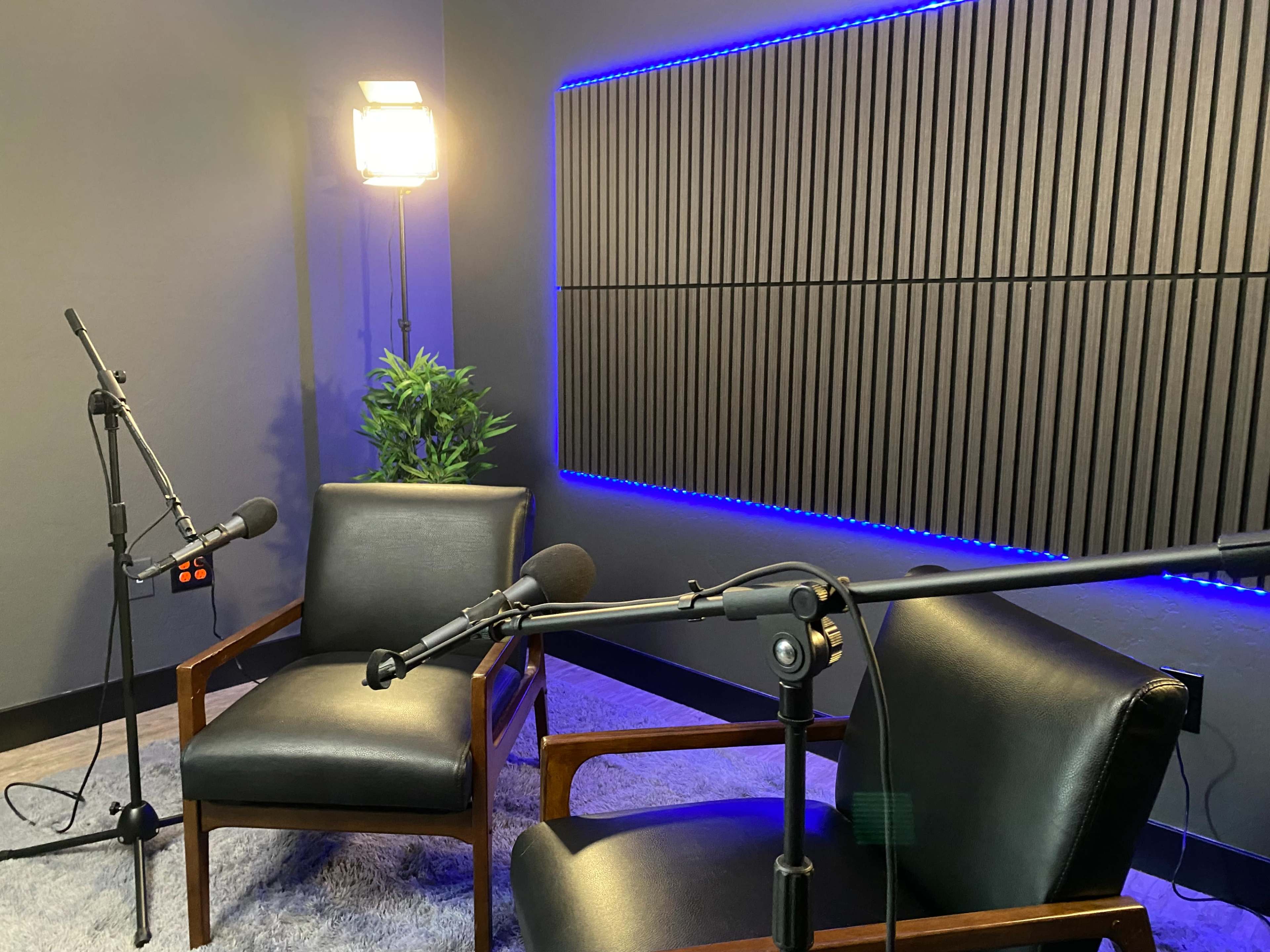 A recording studio setup features two black leather chairs, microphones on stands, and acoustic paneling illuminated by blue lighting.