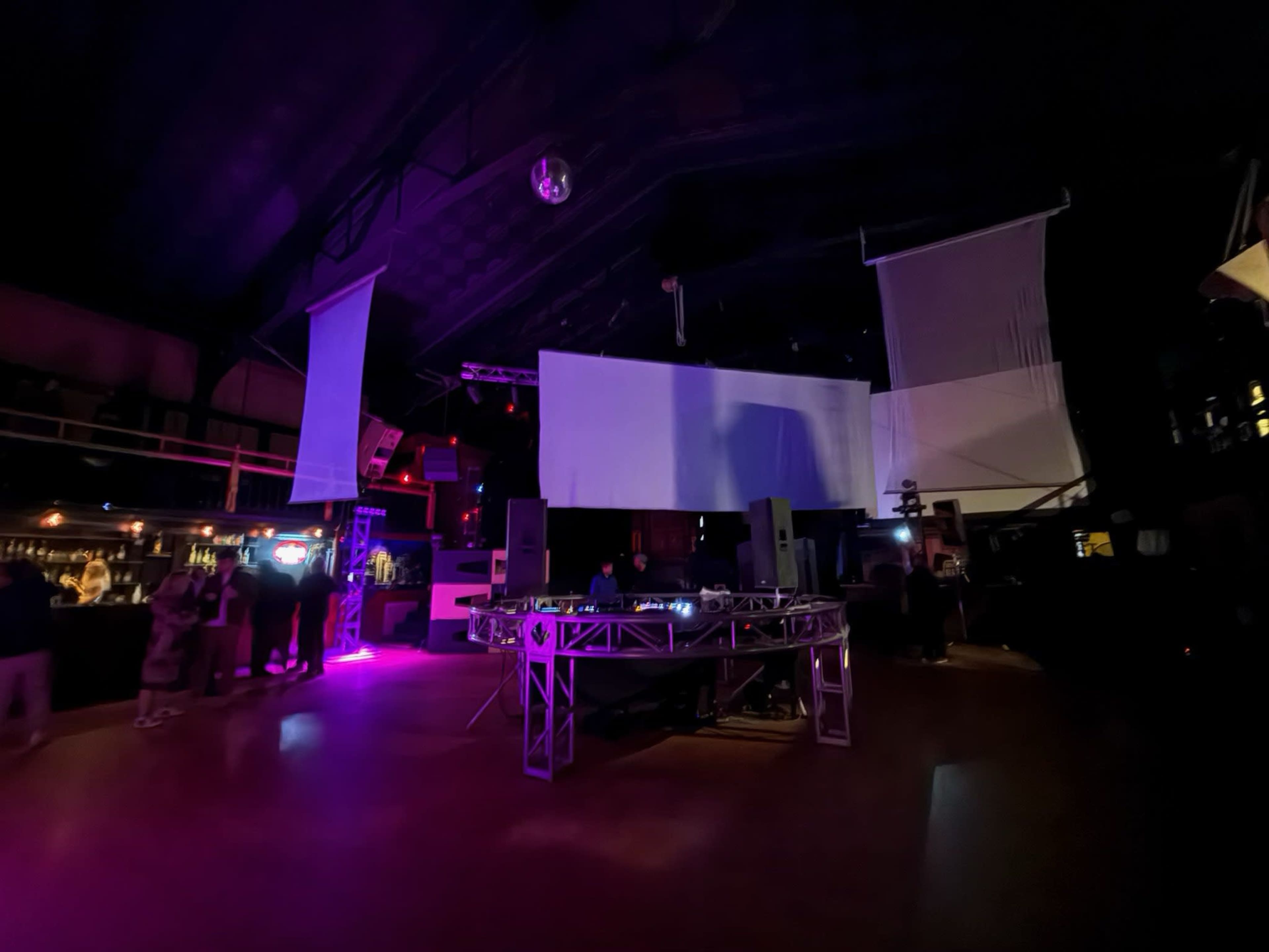 A dimly lit event space features a DJ setup on a raised platform with large speakers, surrounded by colorful lighting and scattered patrons.