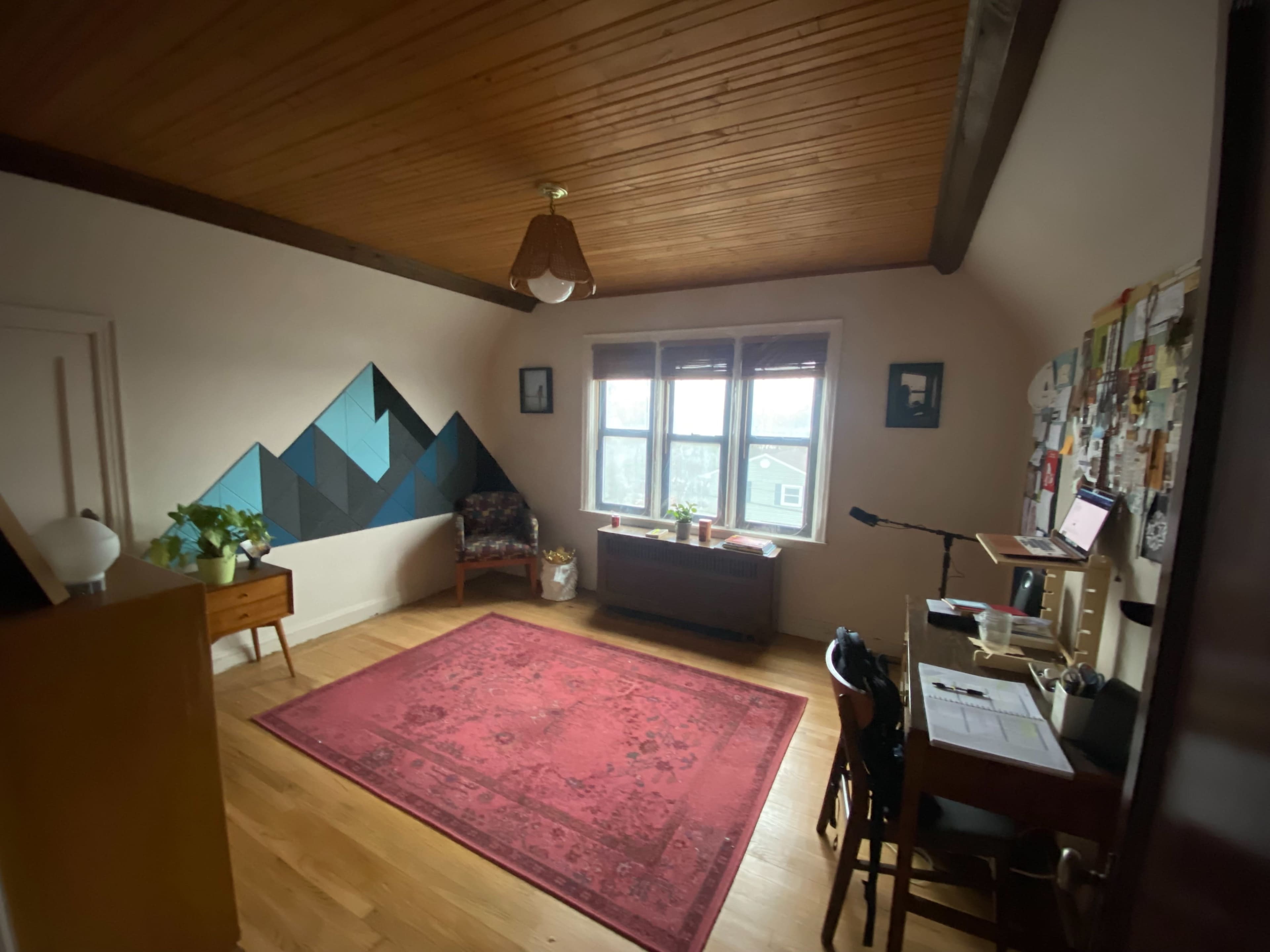 A spacious room with wooden beams on the ceiling, a patterned rug on the floor, a desk with a laptop, and a small seating area by the windows.