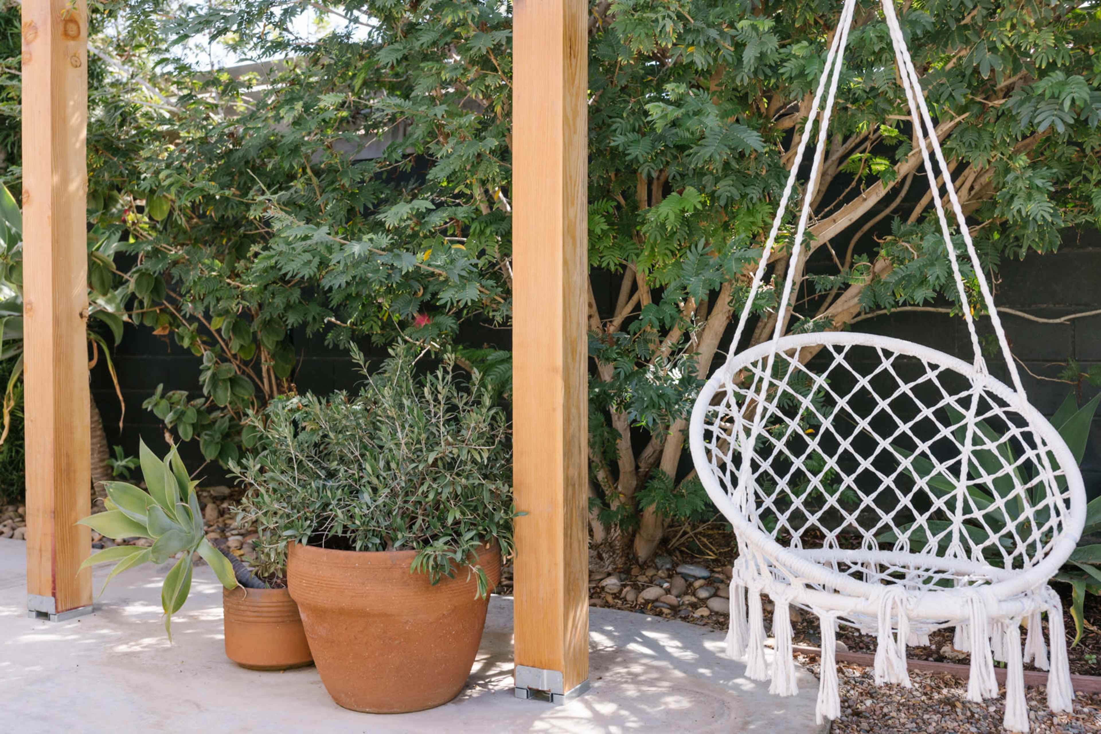 A macramé hanging chair is positioned beside potted plants and wooden posts in a landscaped outdoor area.