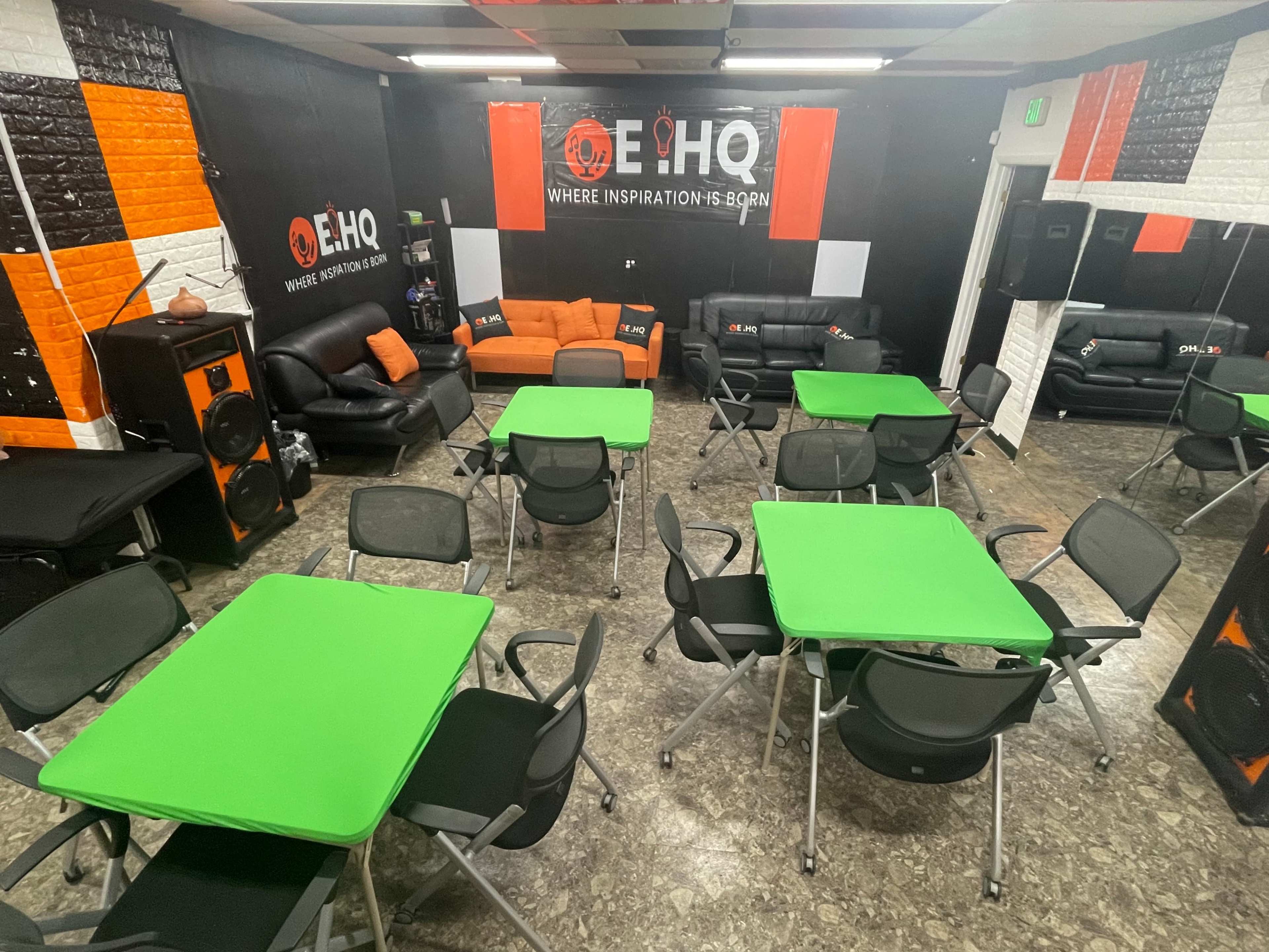 A spacious room arranged with several green tables and black chairs, featuring a lounge area with orange and black seating against a branded wall.