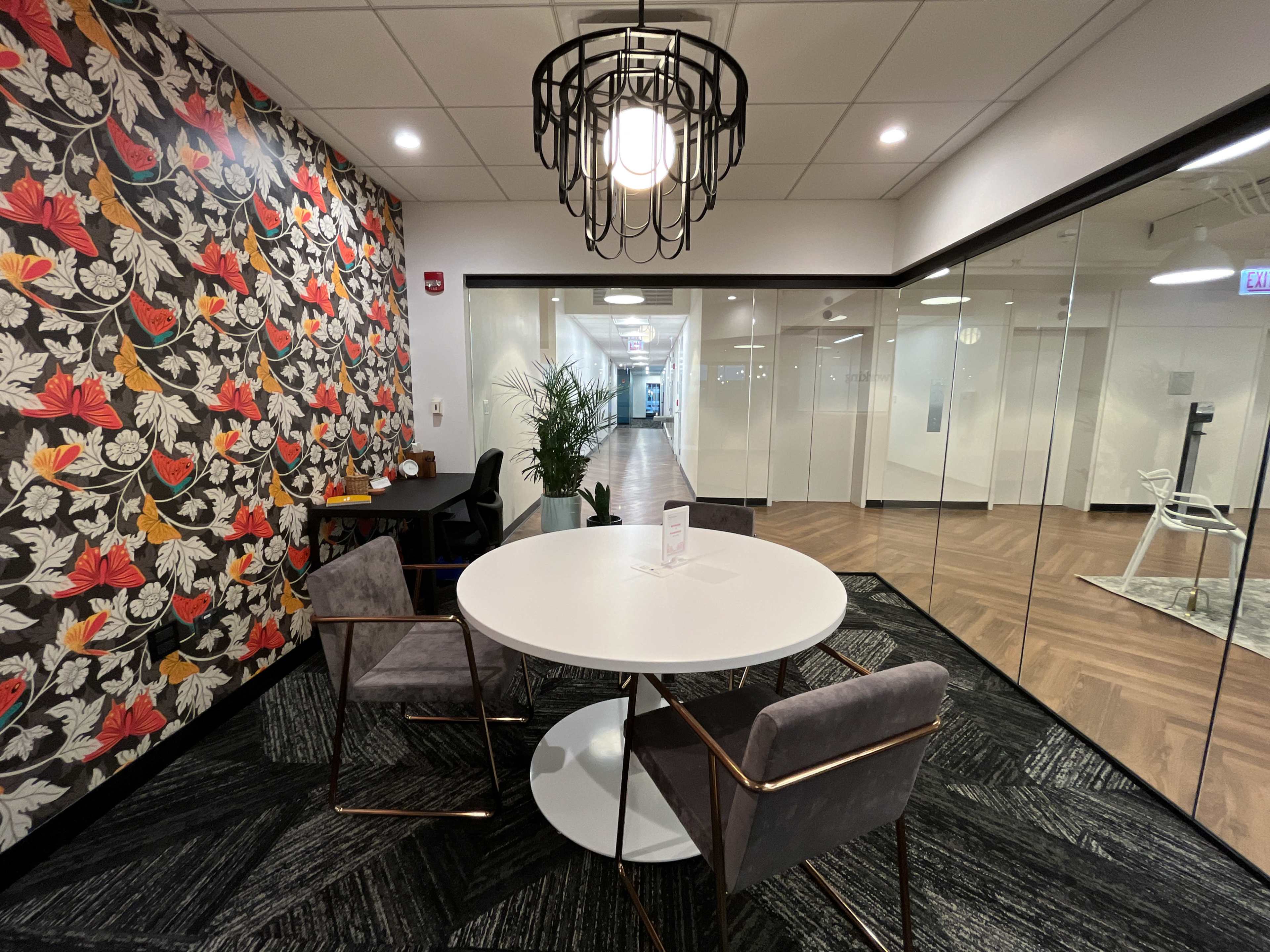 The image shows a modern office space featuring a round white table with two chairs, surrounded by floral wallpaper and glass walls leading to a hallway.