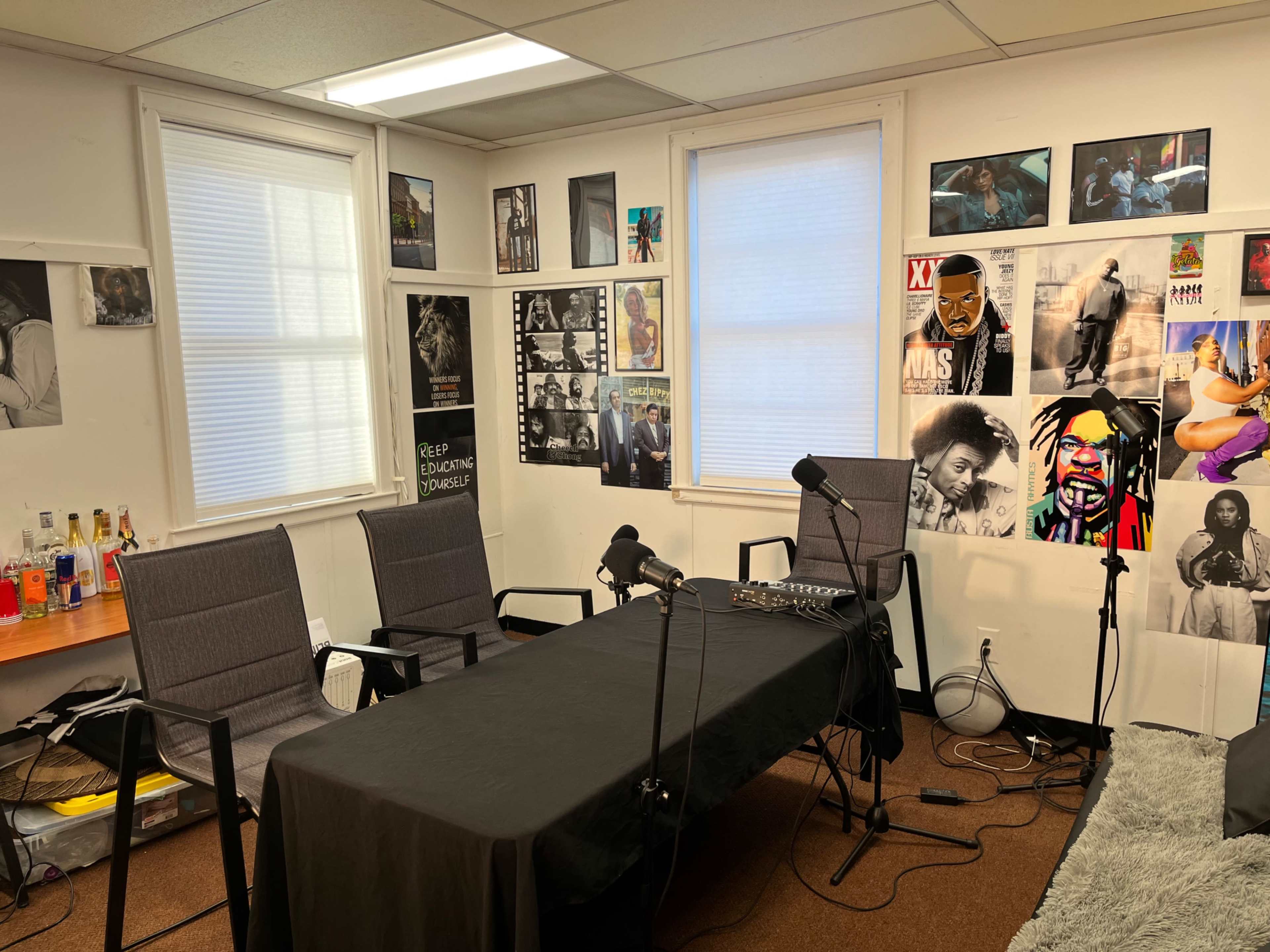 A small room set up for a recording or podcast, featuring a black table, three chairs, microphones, and various photographs on the walls.