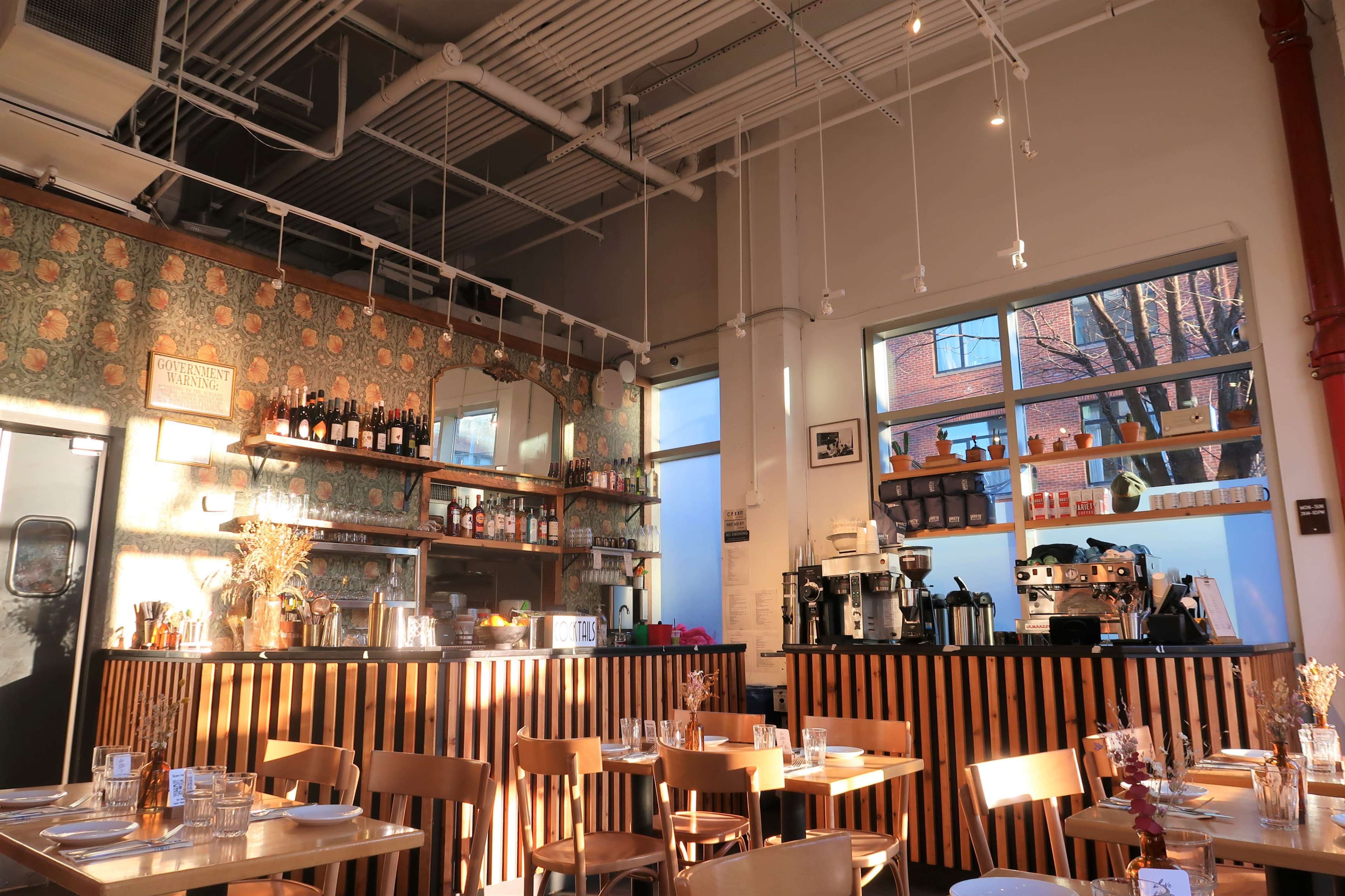 A coffee shop interior with wooden tables, a bar area lined with bottles and a coffee machine, and large windows letting in natural light.