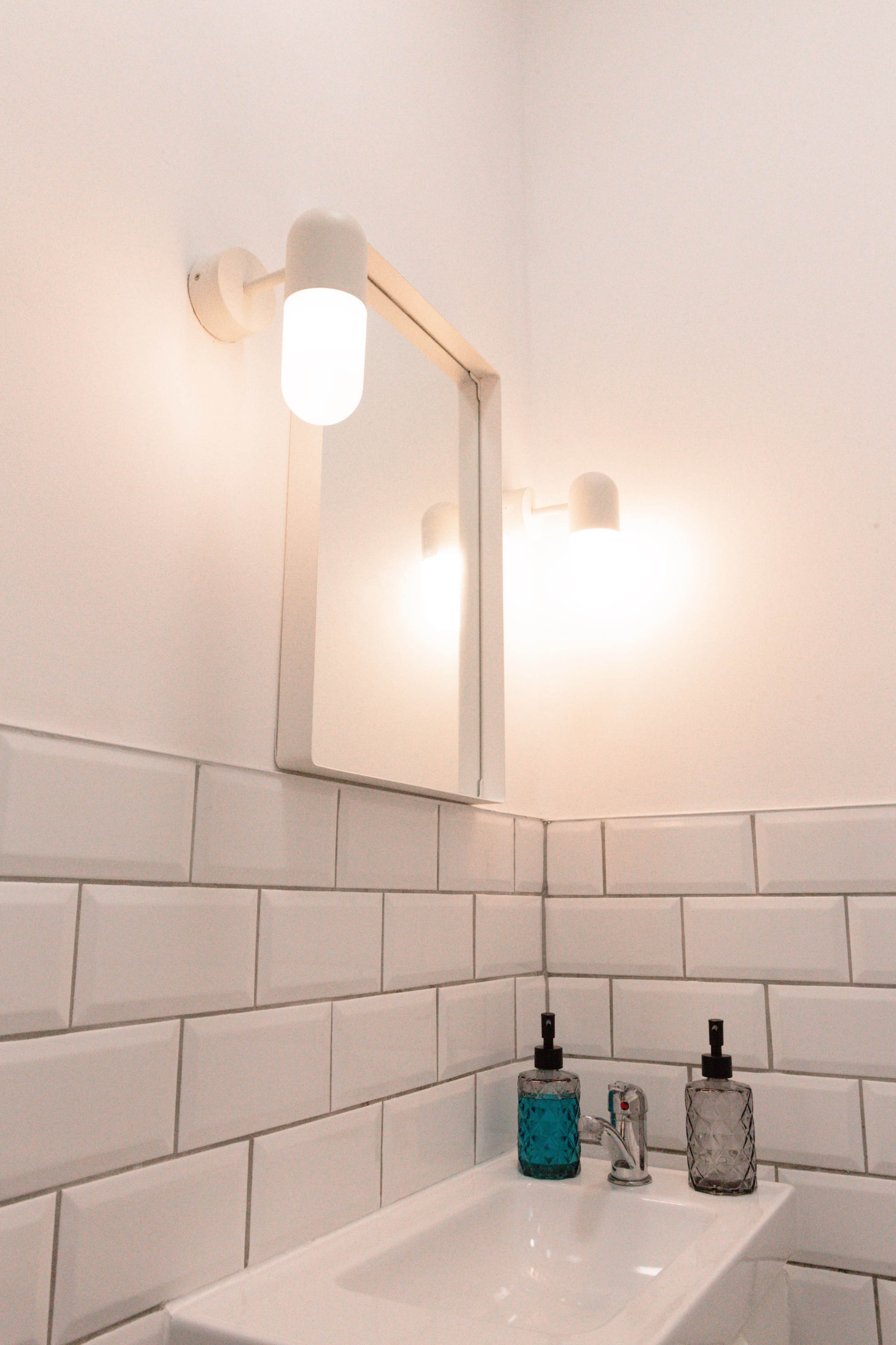 A modern bathroom features a white tiled wall, a rectangular mirror with an overhead light, and two bottles next to a sink.