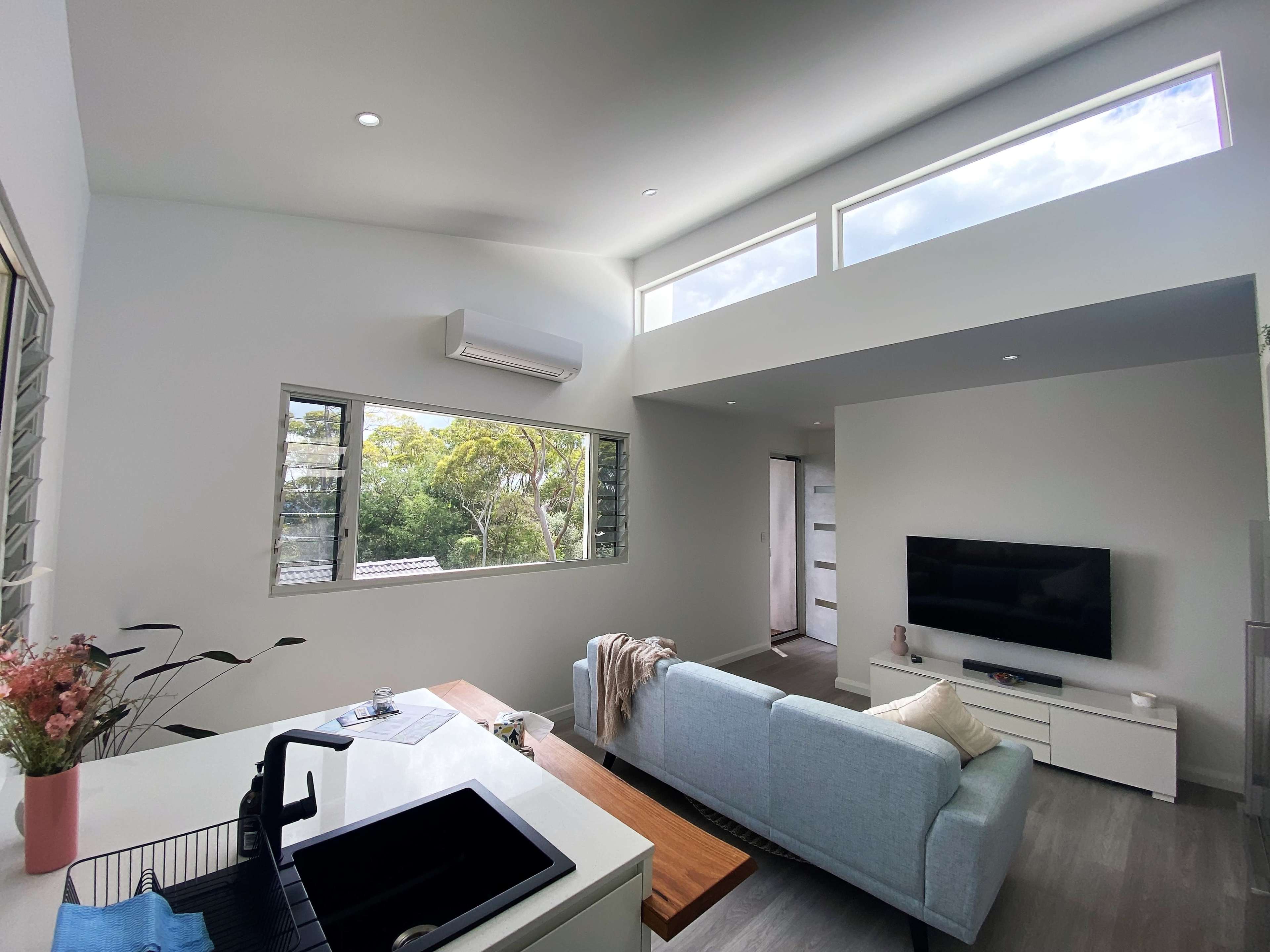 A modern living room with large windows, a light-colored sofa, a television mounted on the wall, and a kitchen area featuring a sink and countertop.