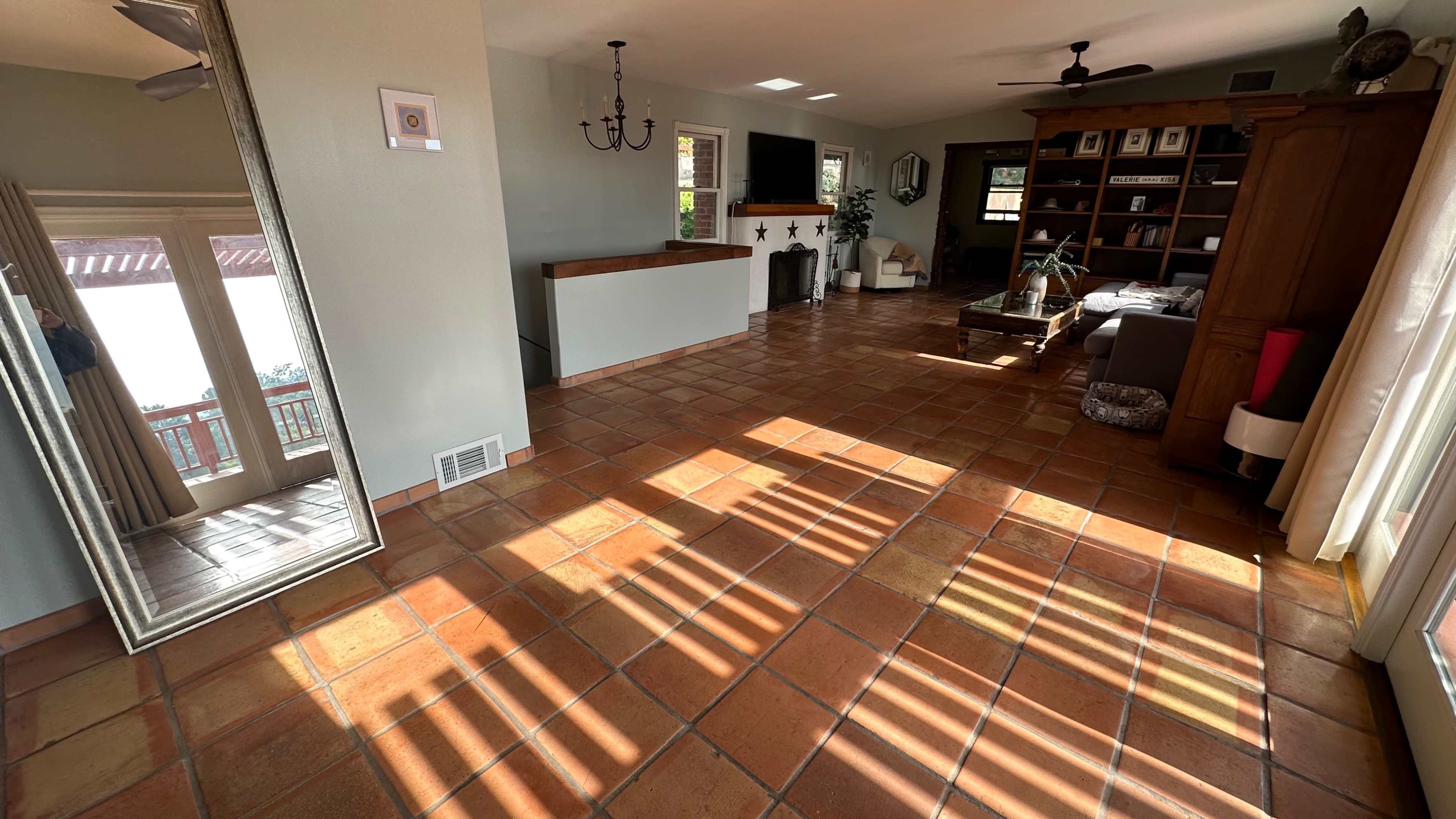 A spacious living room with terracotta tile flooring, a large mirror, and sunlight casting striped shadows on the floor.