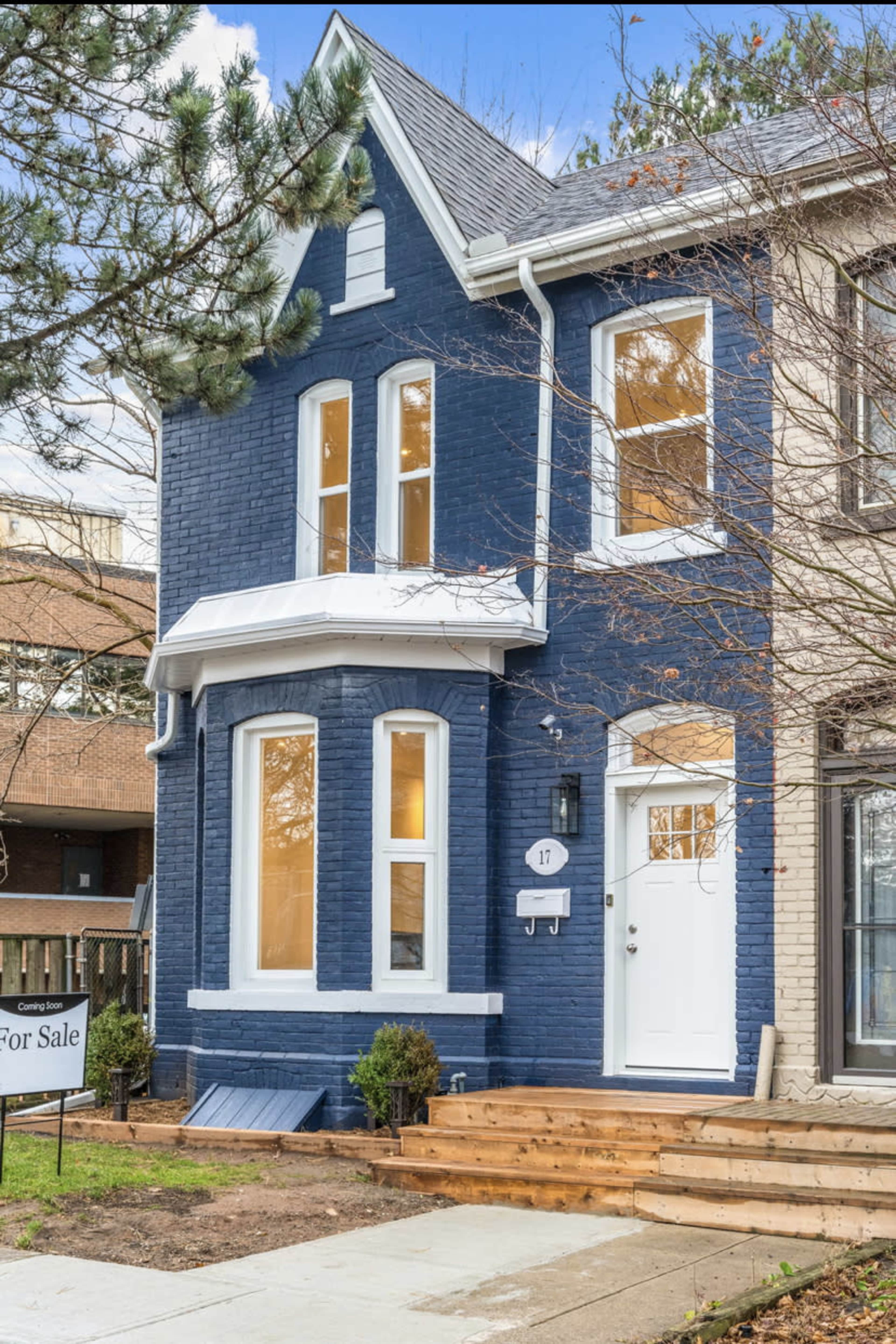 A two-story blue brick house with large windows and a wooden staircase leading to the entrance, marked with a "For Sale" sign.