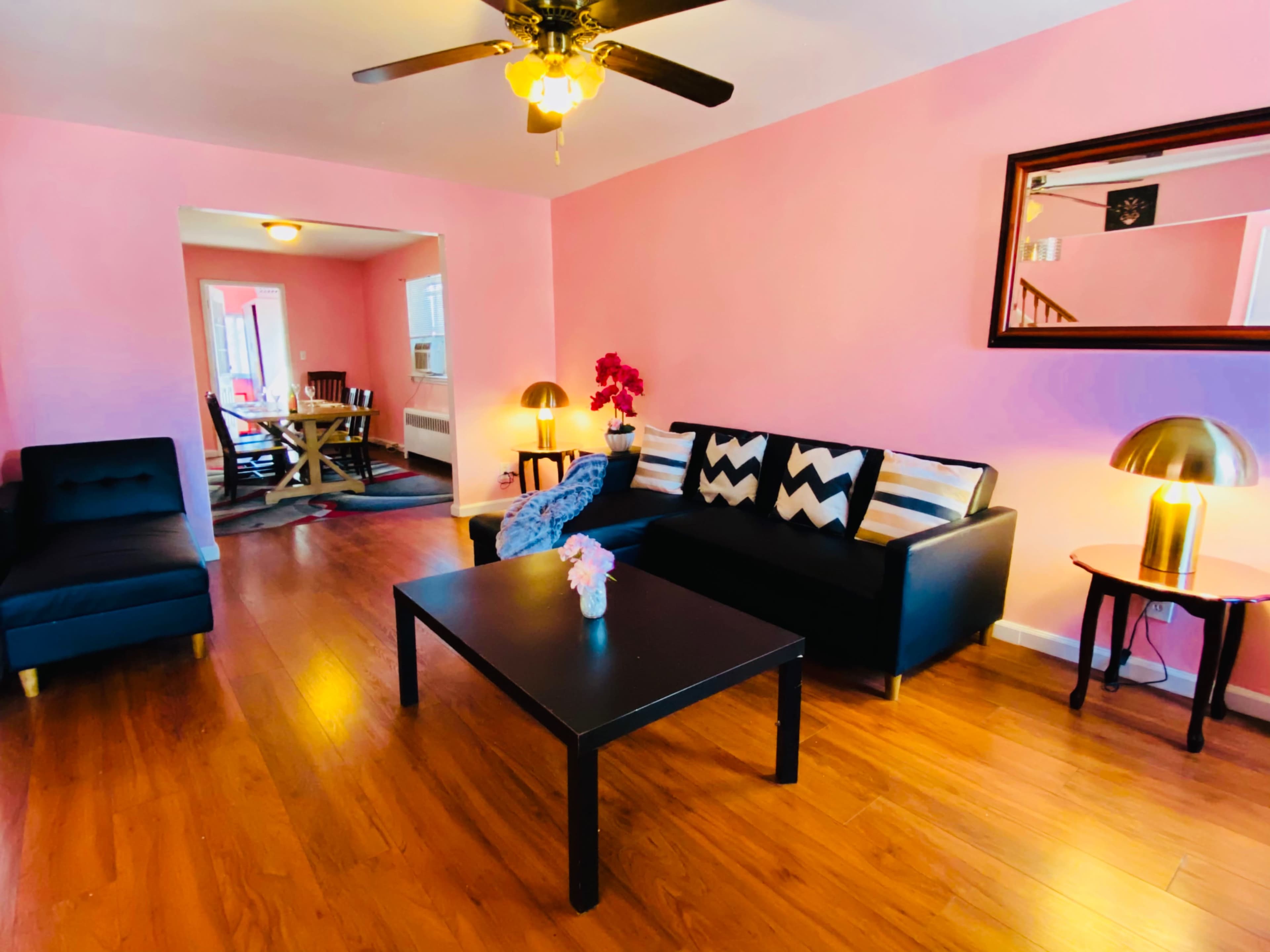 A living room with pink walls, a black sofa and coffee table, and a dining area visible in the background.