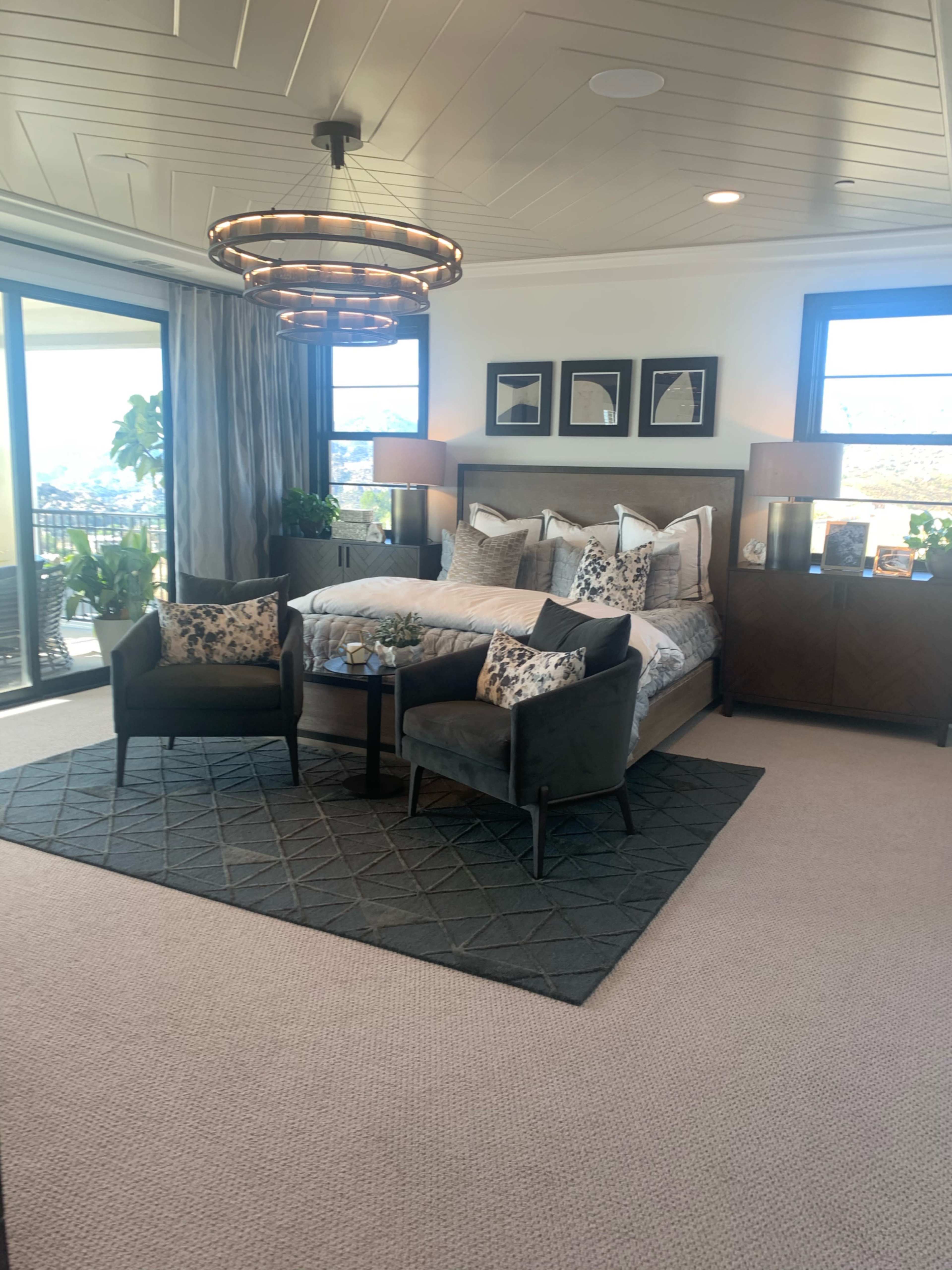 The image shows a modern bedroom with a large bed, two upholstered chairs, a round coffee table, and a large window that offers a view of the outdoors.