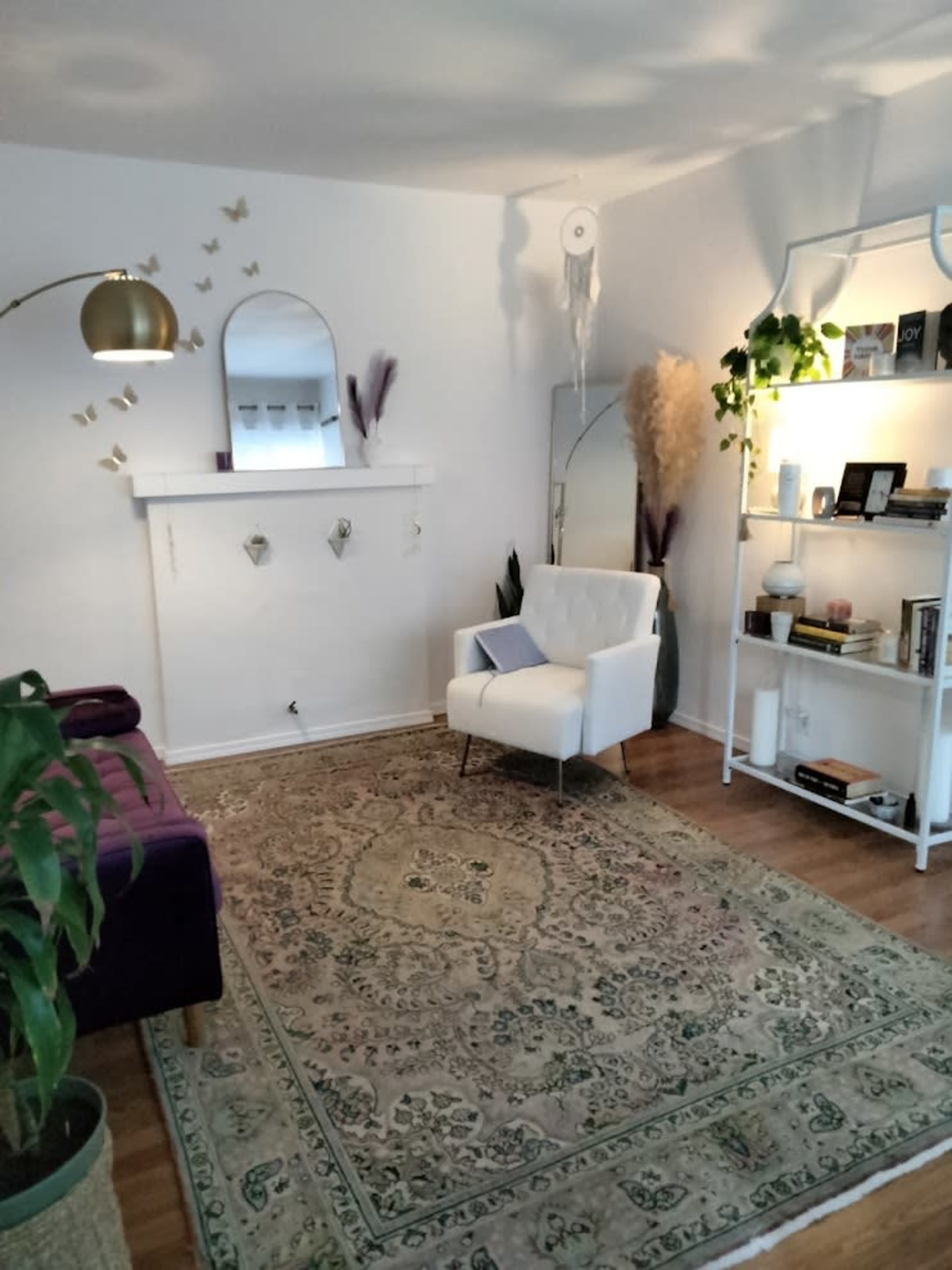 The image shows a living room with a white armchair, a purple sofa, a decorative rug, wall decor, and a bookshelf filled with books and plants.