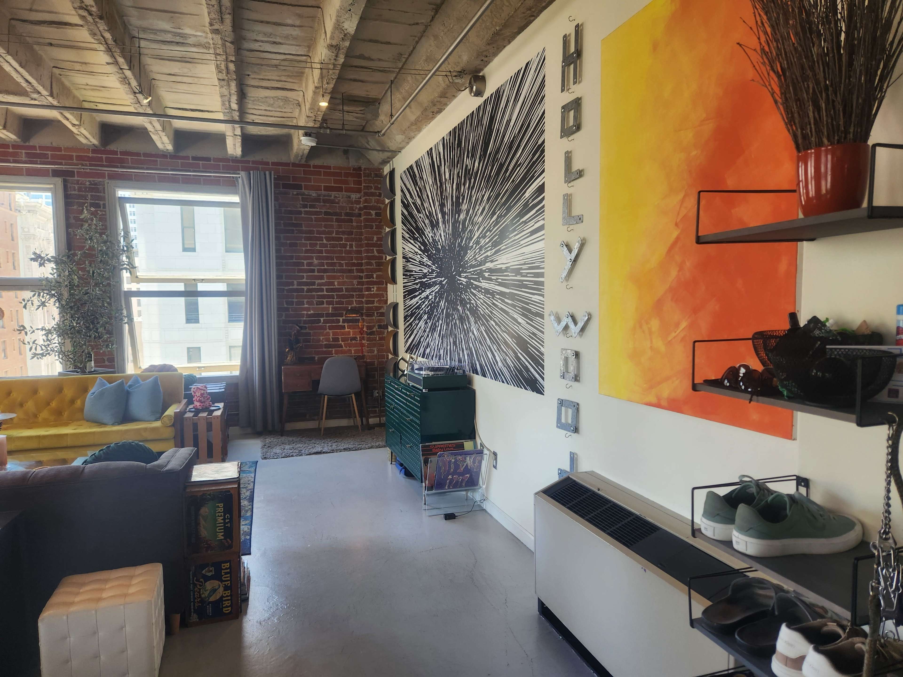 The image shows a modern urban living space featuring a yellow sofa, a wall with a large graphic art piece, and a decorative shelf with shoes and plants.