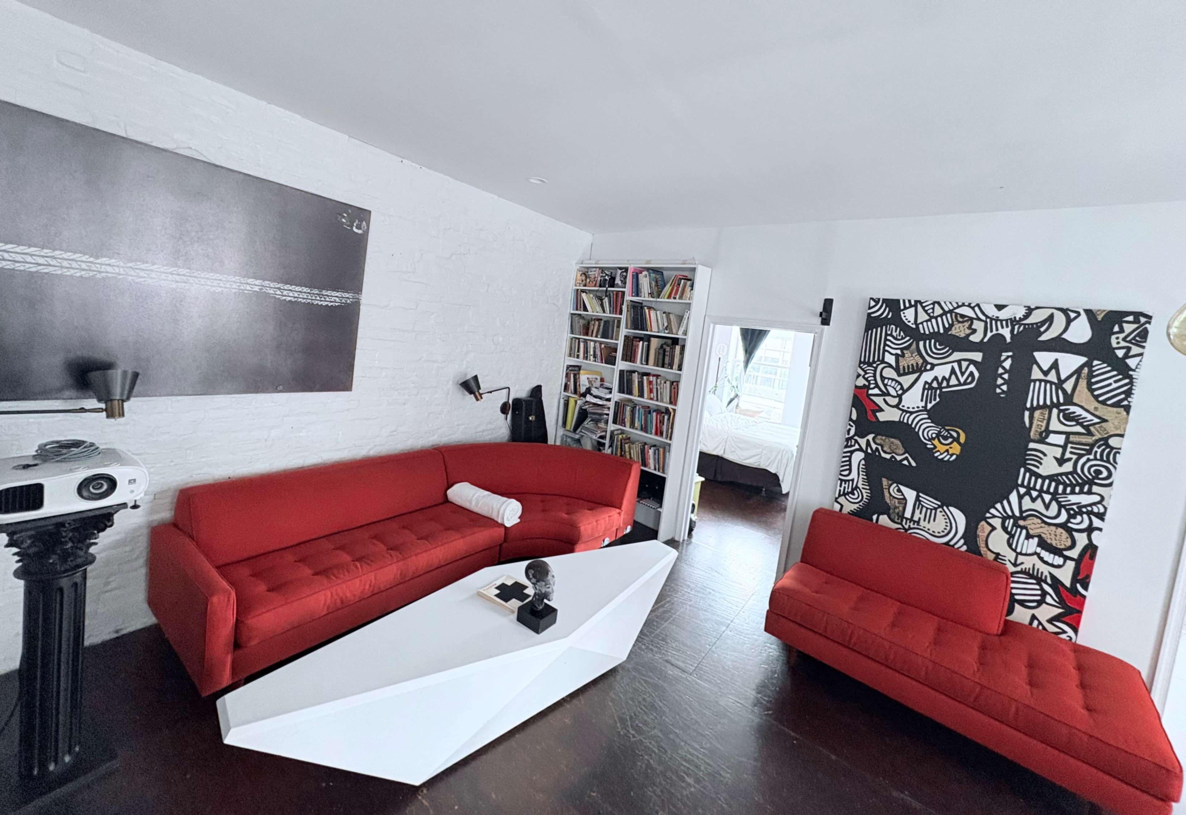 The image shows a modern living room featuring two red sofas, a white coffee table, a black and white artwork on the wall, and a bookshelf filled with books.