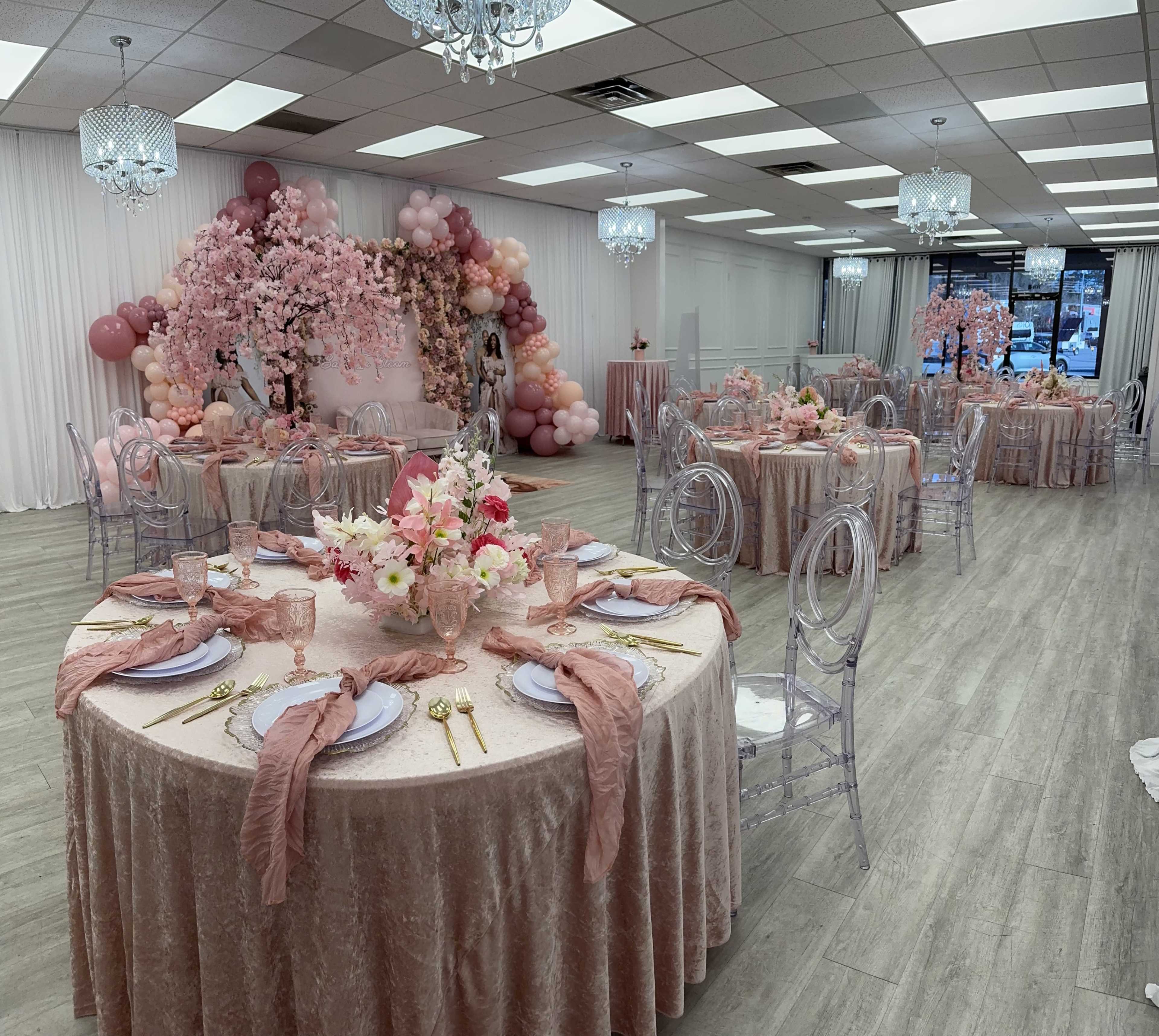 The image shows a banquet hall decorated for an event, featuring tables with pink tablecloths, floral centerpieces, and a backdrop of balloons and cherry blossoms.