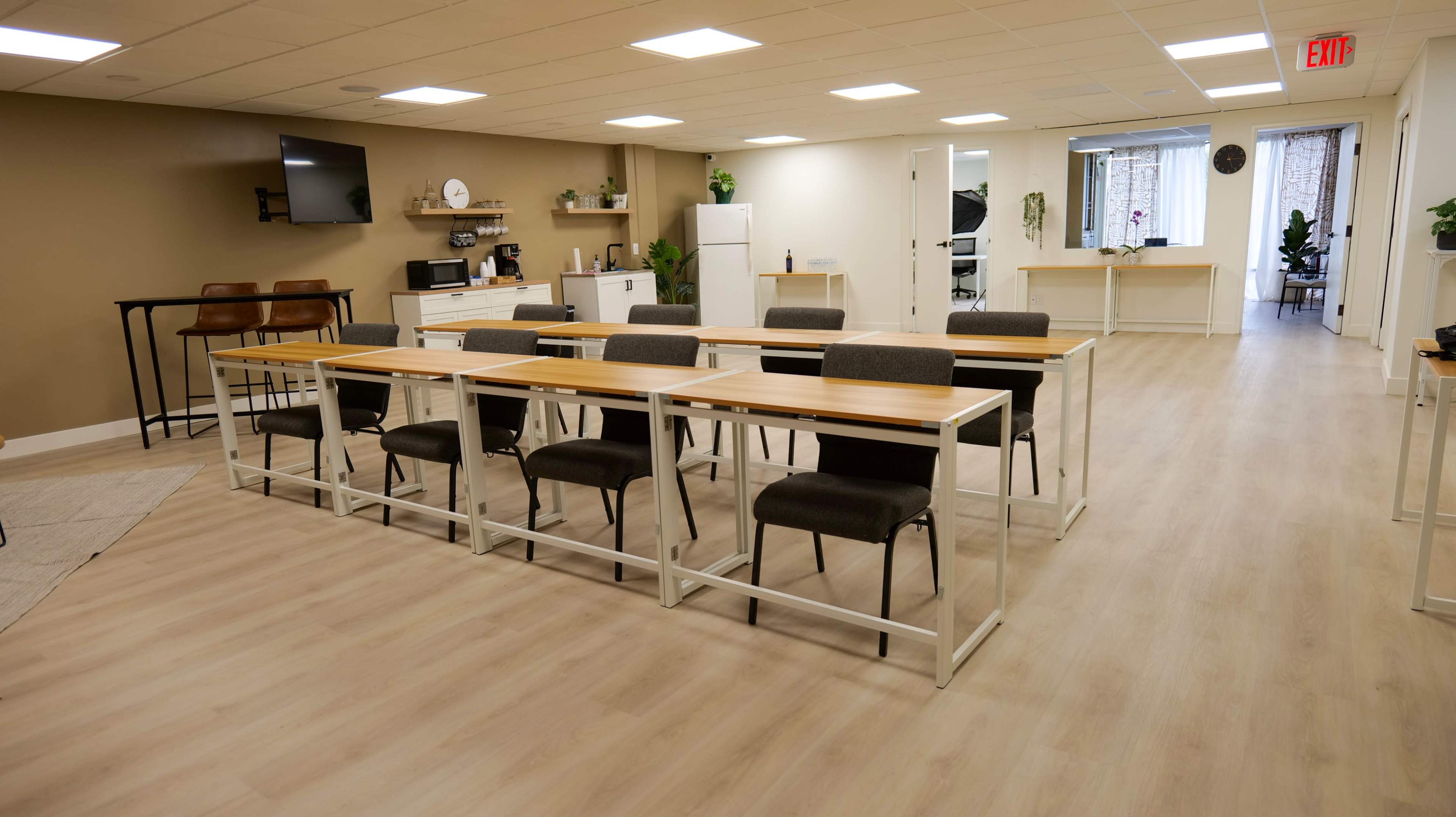 A modern, bright room features several tables with chairs arranged in a row, along with kitchen appliances and plants lining the walls.