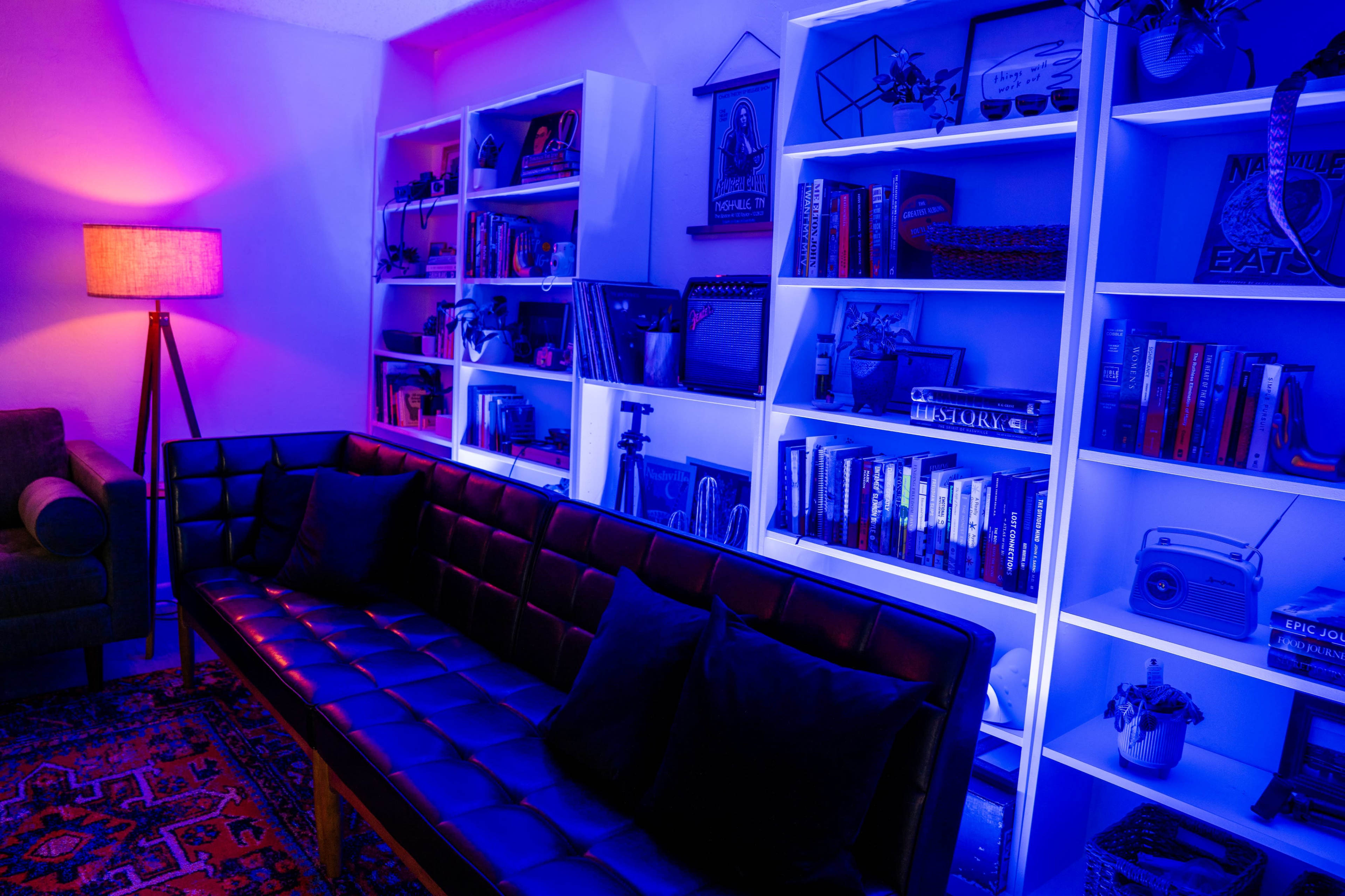 A modern living room features a black leather sofa, a standing lamp, and white bookshelves illuminated with blue and purple lighting, filled with books and various decorative items.