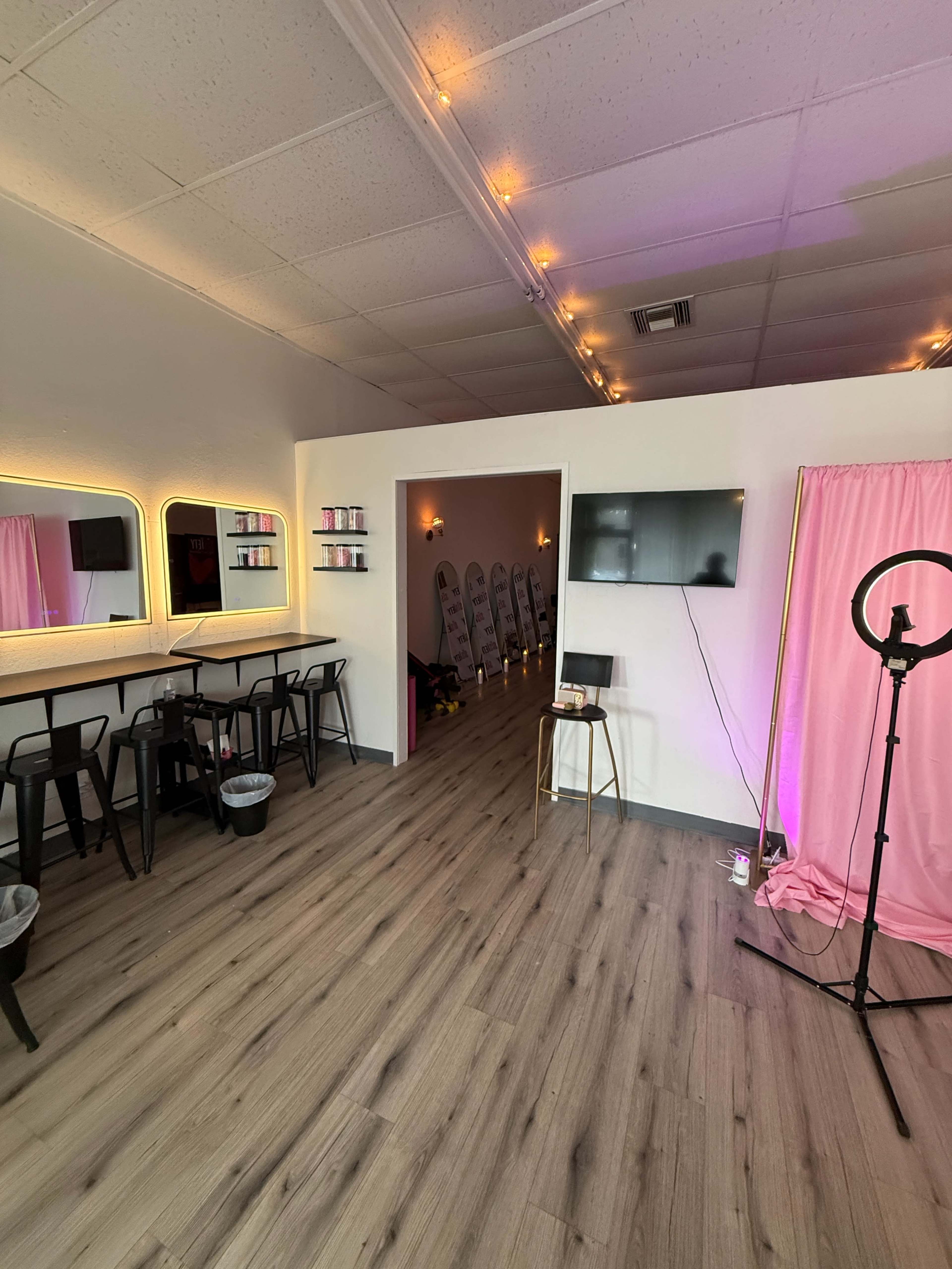 The image shows a stylish, well-lit interior of a beauty studio featuring a seating area with high stools, a segment with illuminated mirrors, and a corridor leading to a room with several hair styling stations.