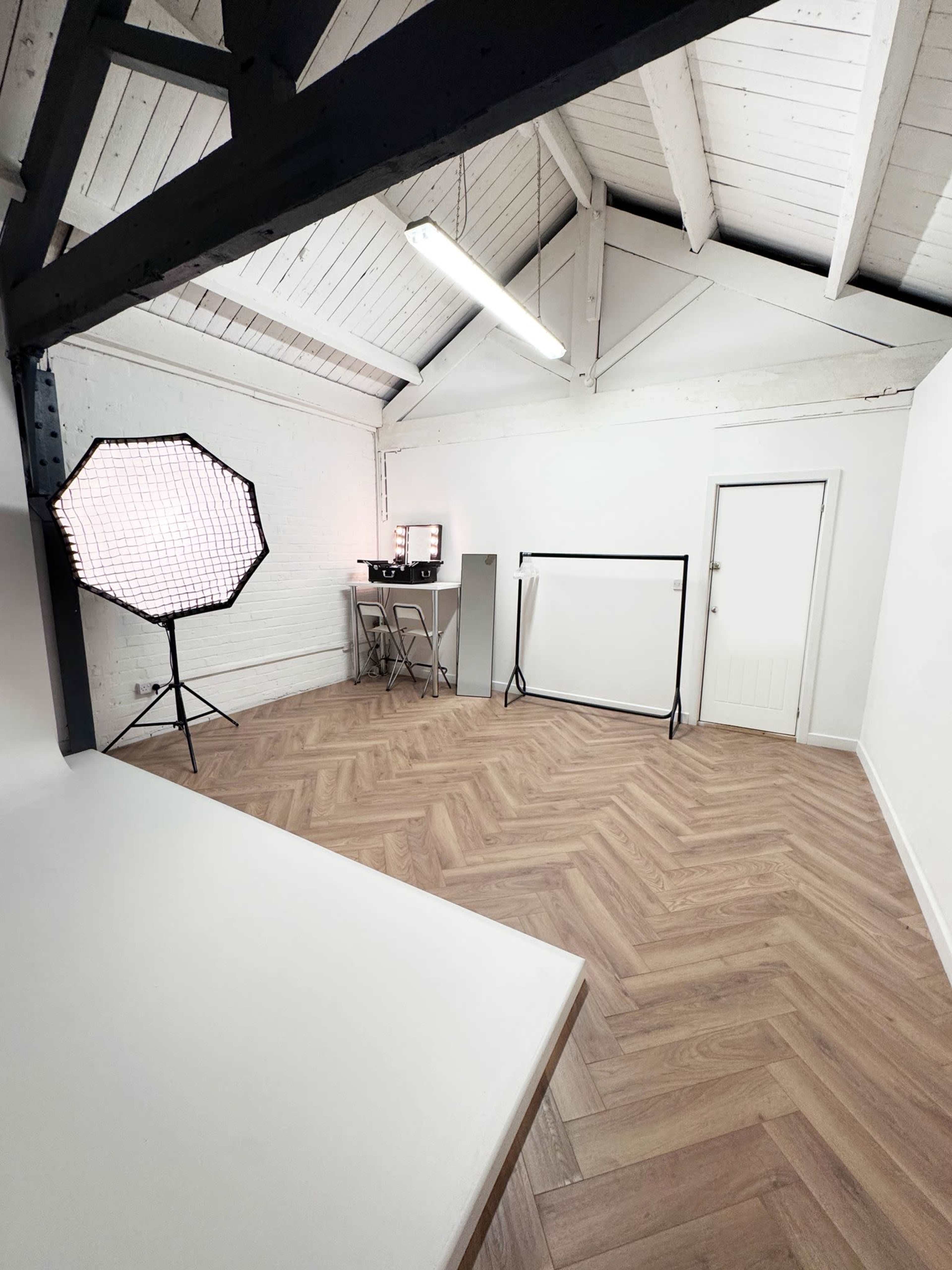 The image shows a spacious, well-lit photo studio with a herringbone wooden floor, equipped with lighting setup, a small table with equipment, and a backdrop stand.