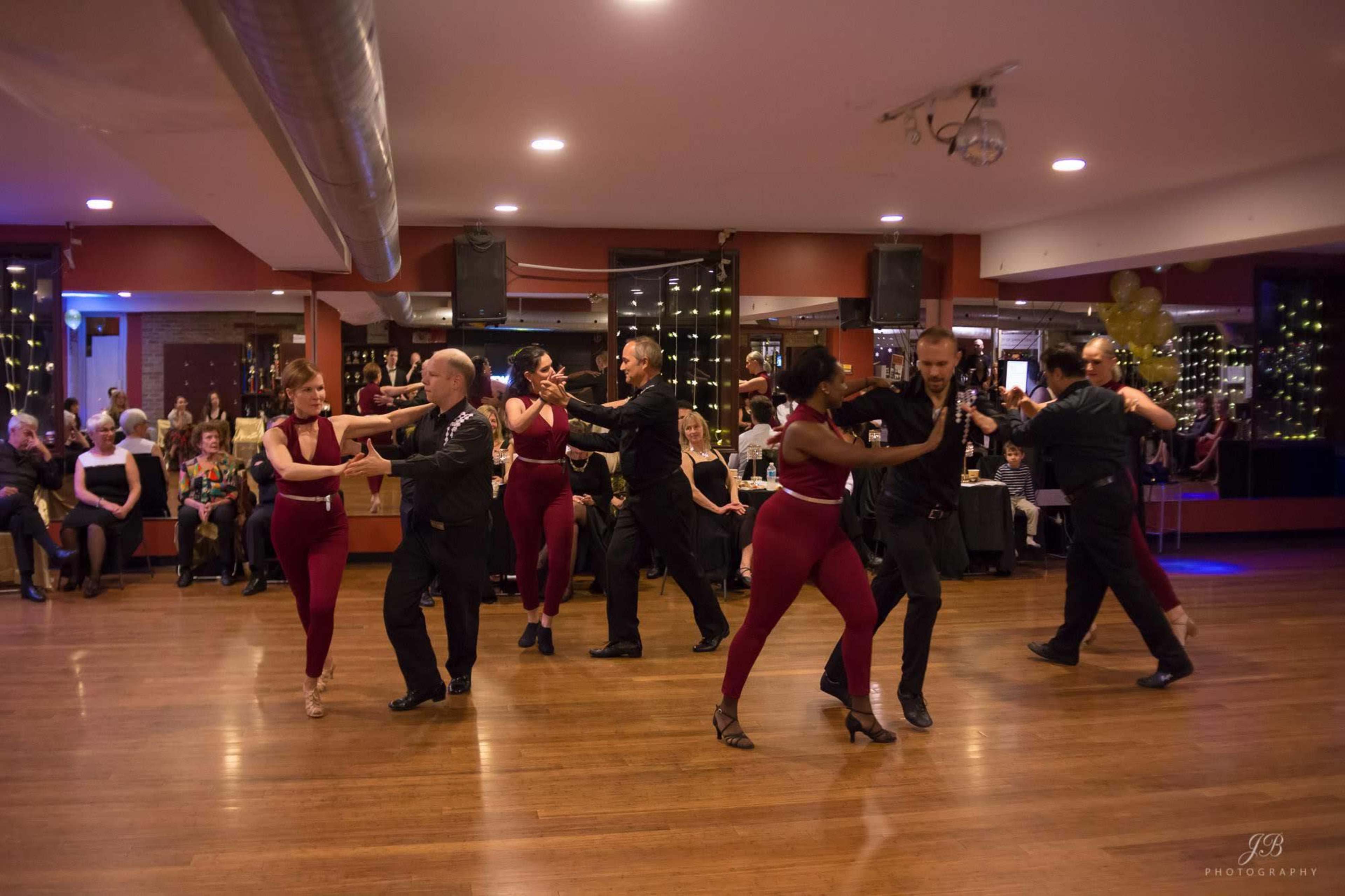 A group of six dancers performs a tango on a wooden dance floor, while an audience watches from seated positions in the background.
