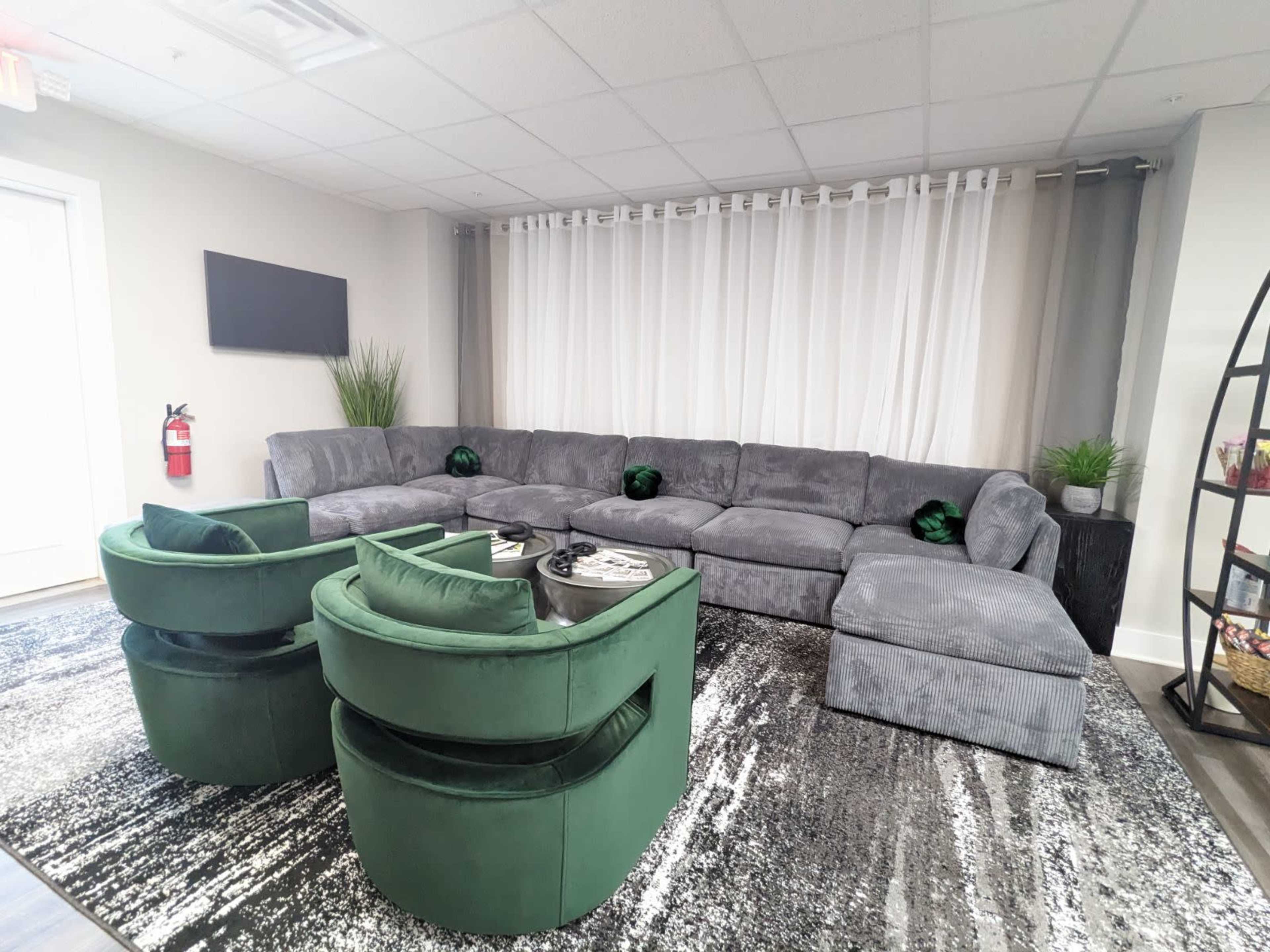 A modern lounge area featuring a large gray sectional sofa, two green velvet chairs, and a black and white patterned rug.