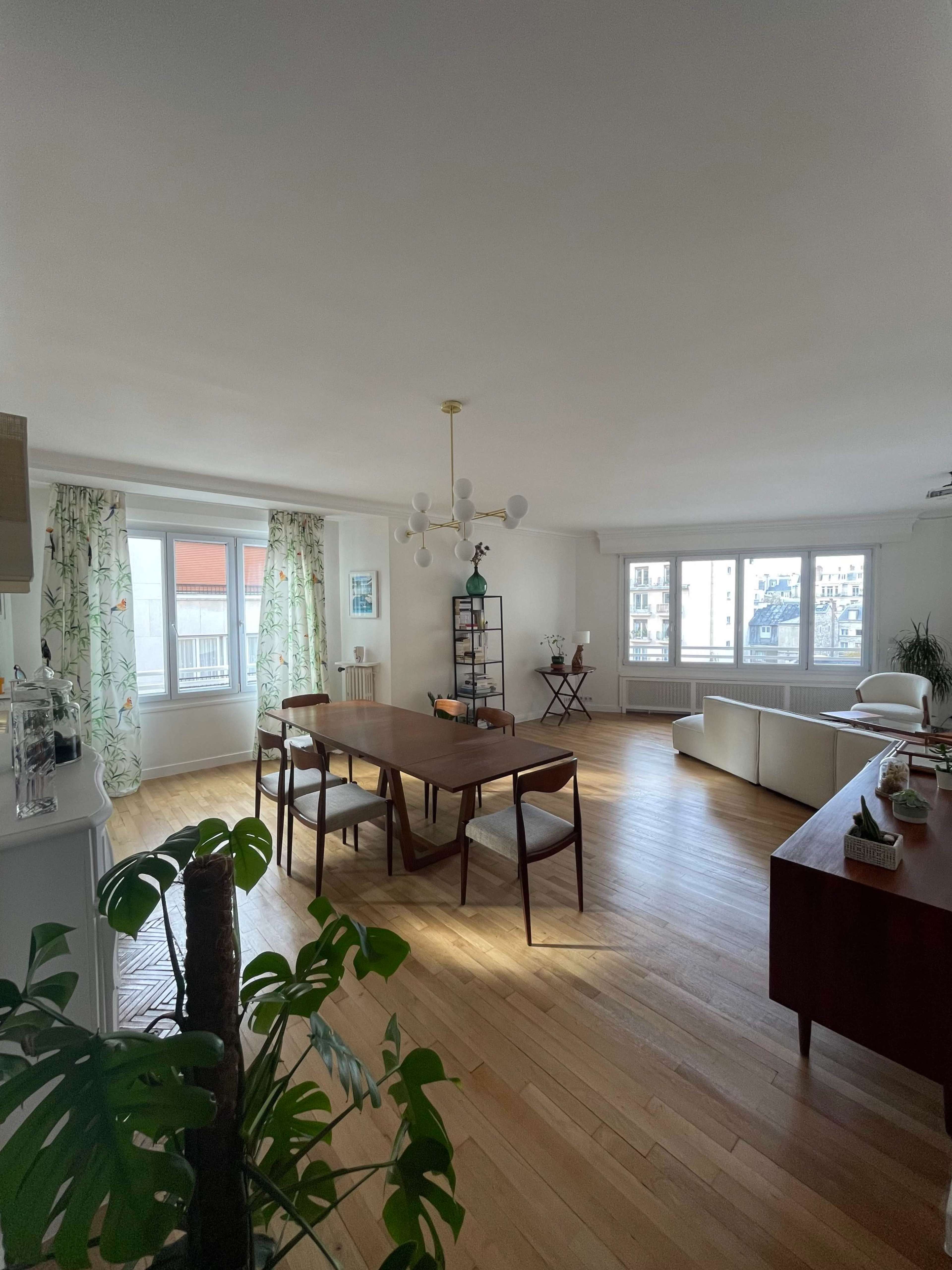 A spacious, bright living area features a dining table, several chairs, a modern light fixture, and large windows with floral curtains.