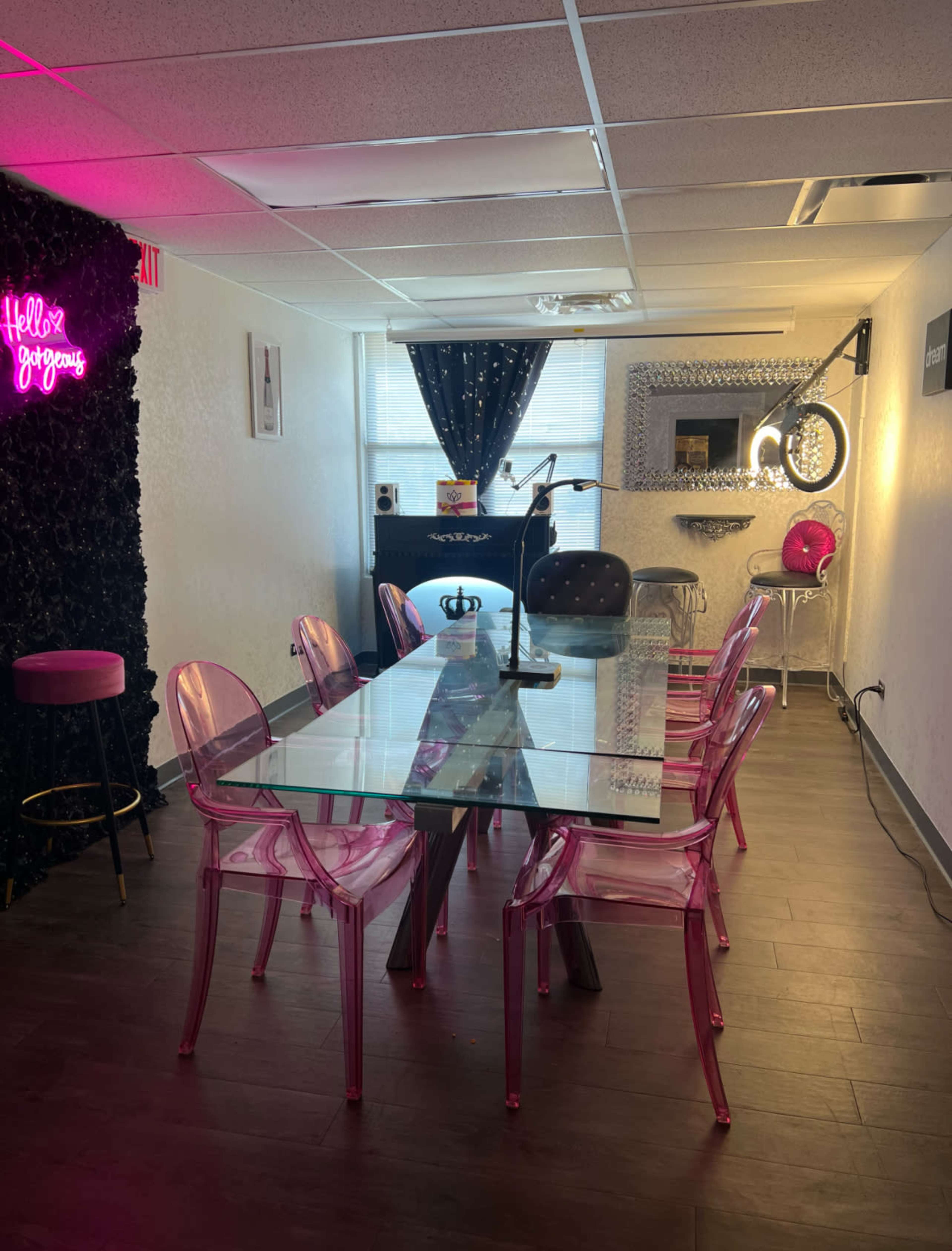 The image shows a modern meeting room with a glass table surrounded by transparent pink chairs, featuring decorative walls and a neon sign.
