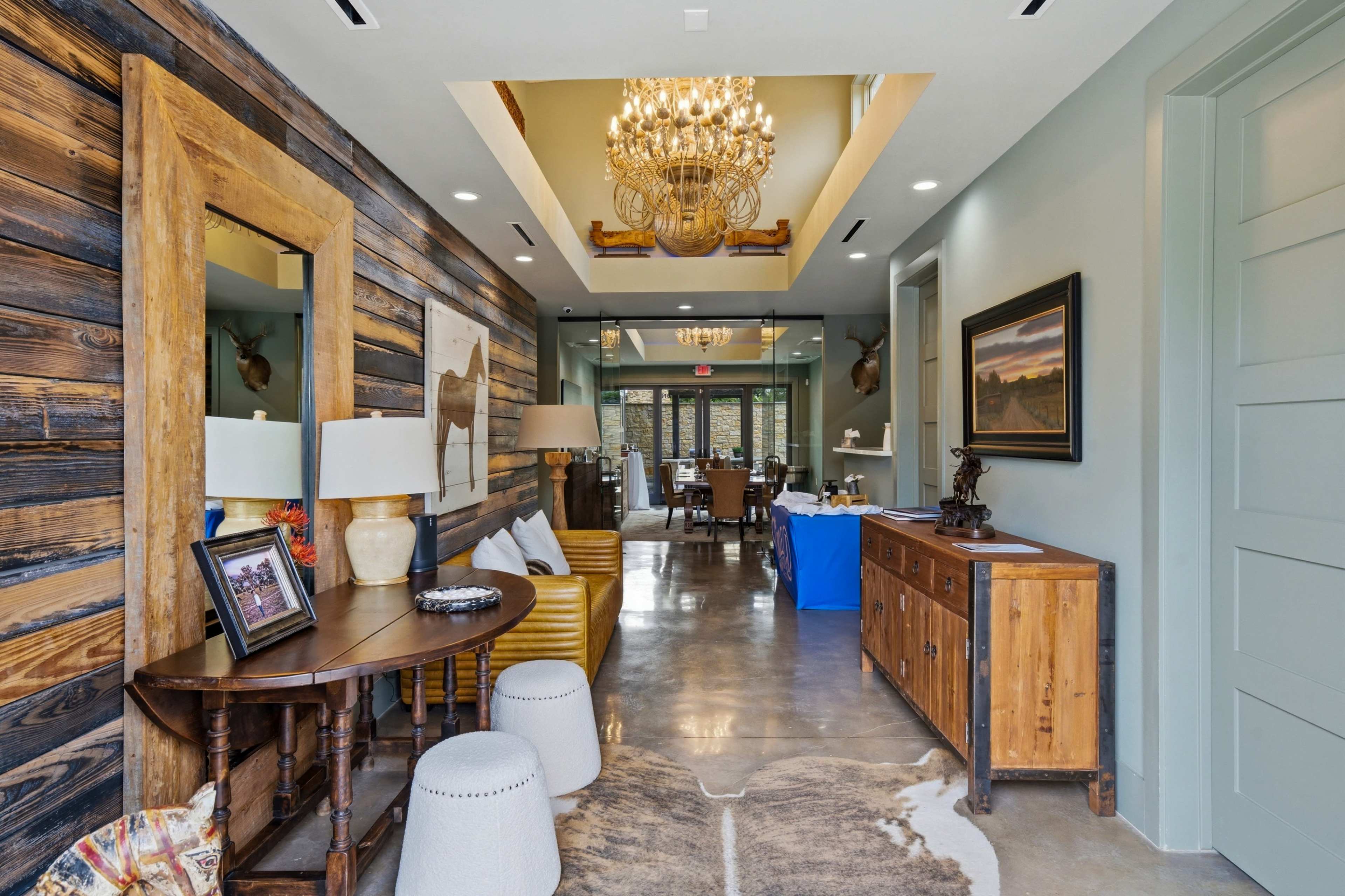The image shows a brightly lit hallway with wood-paneled walls, a decorative mirror, a side table, and a large chandelier, leading into a dining area.