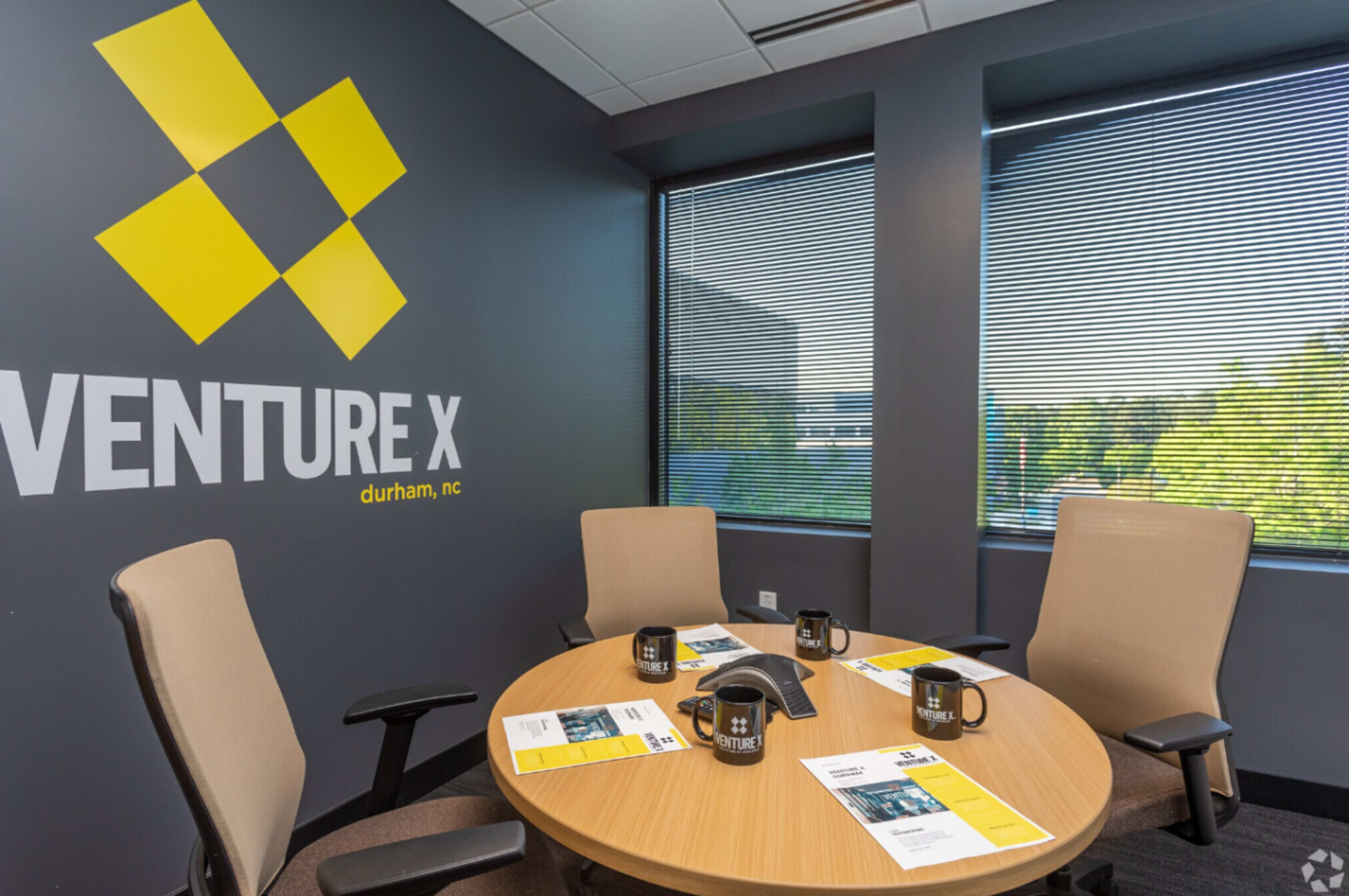 A meeting room at Venture X in Durham, NC, featuring a round wooden table with two chairs and promotional materials on the surface.