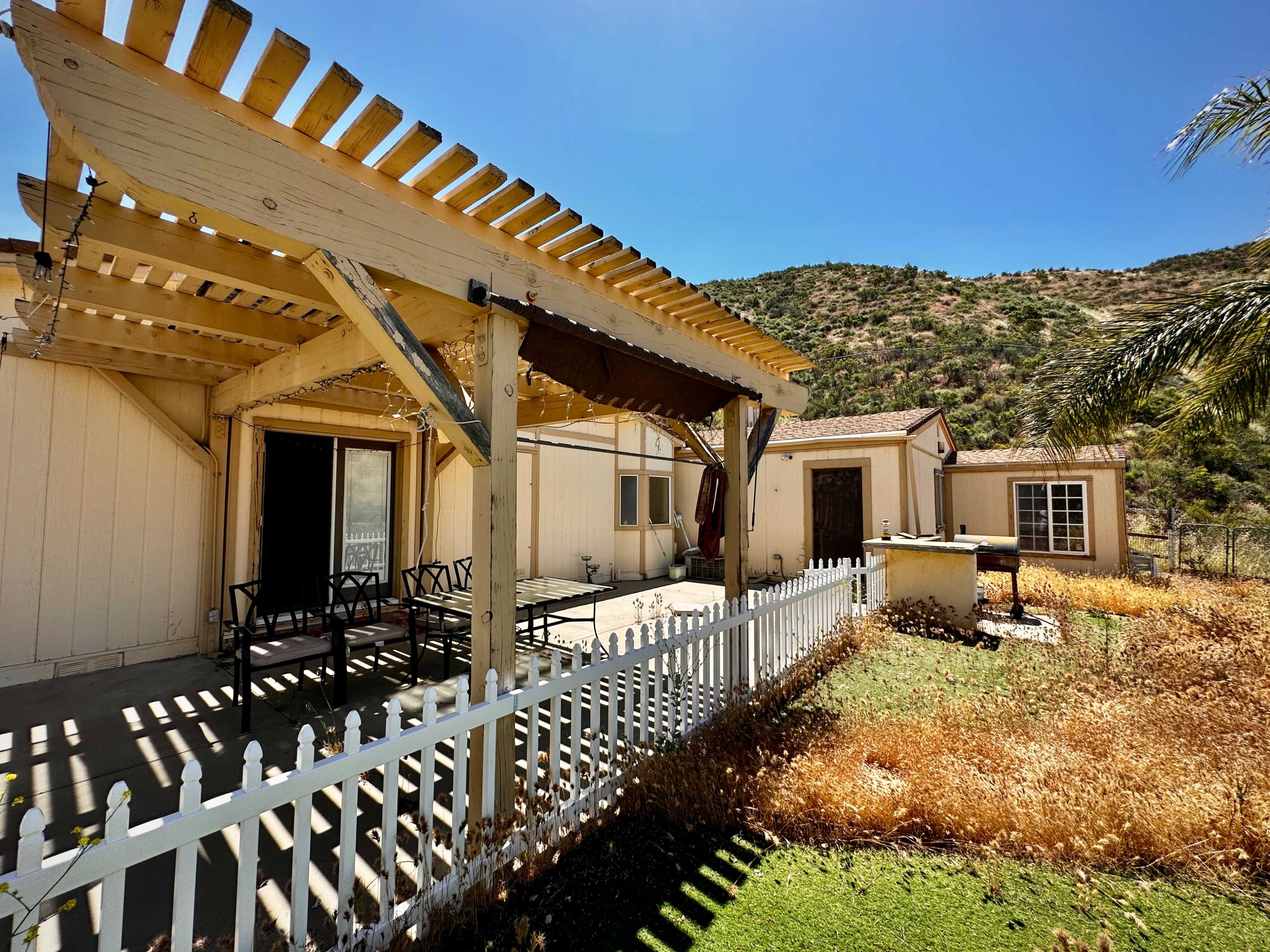 A wooden pergola shades a patio area beside a house surrounded by overgrown grass and hills in the background.
