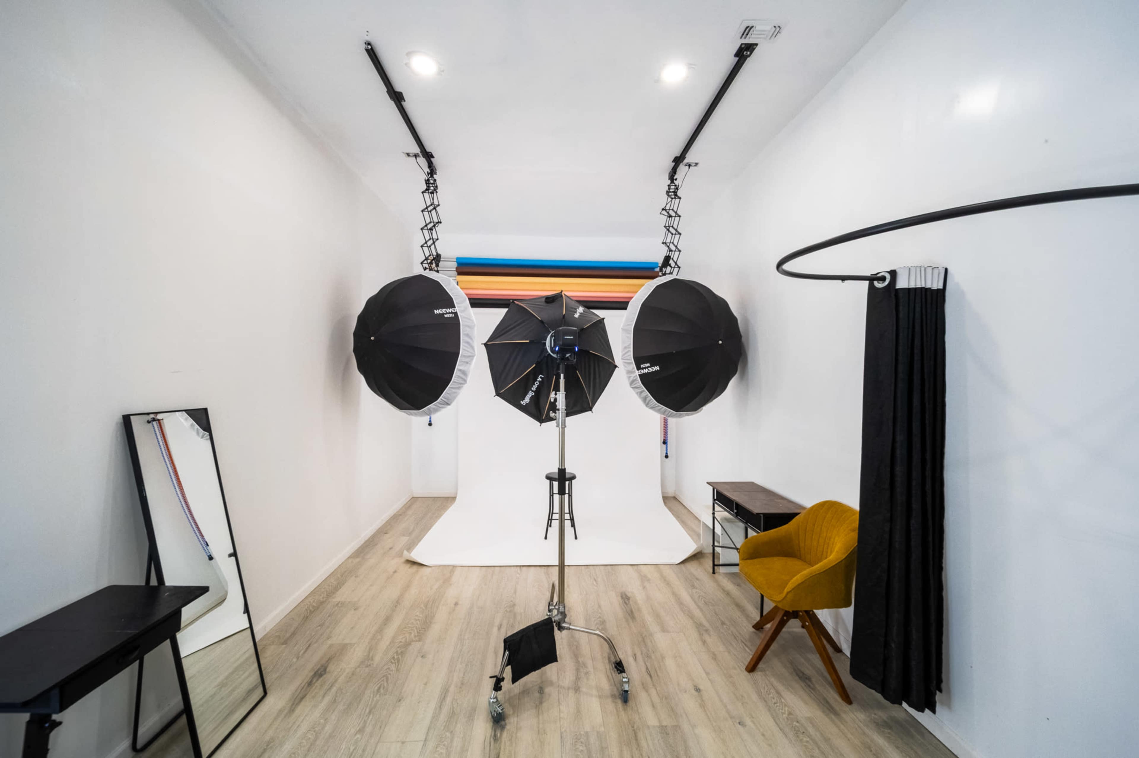The image shows a photography studio with three softbox lights, a white backdrop, and a mirror alongside a black curtain and a yellow chair.