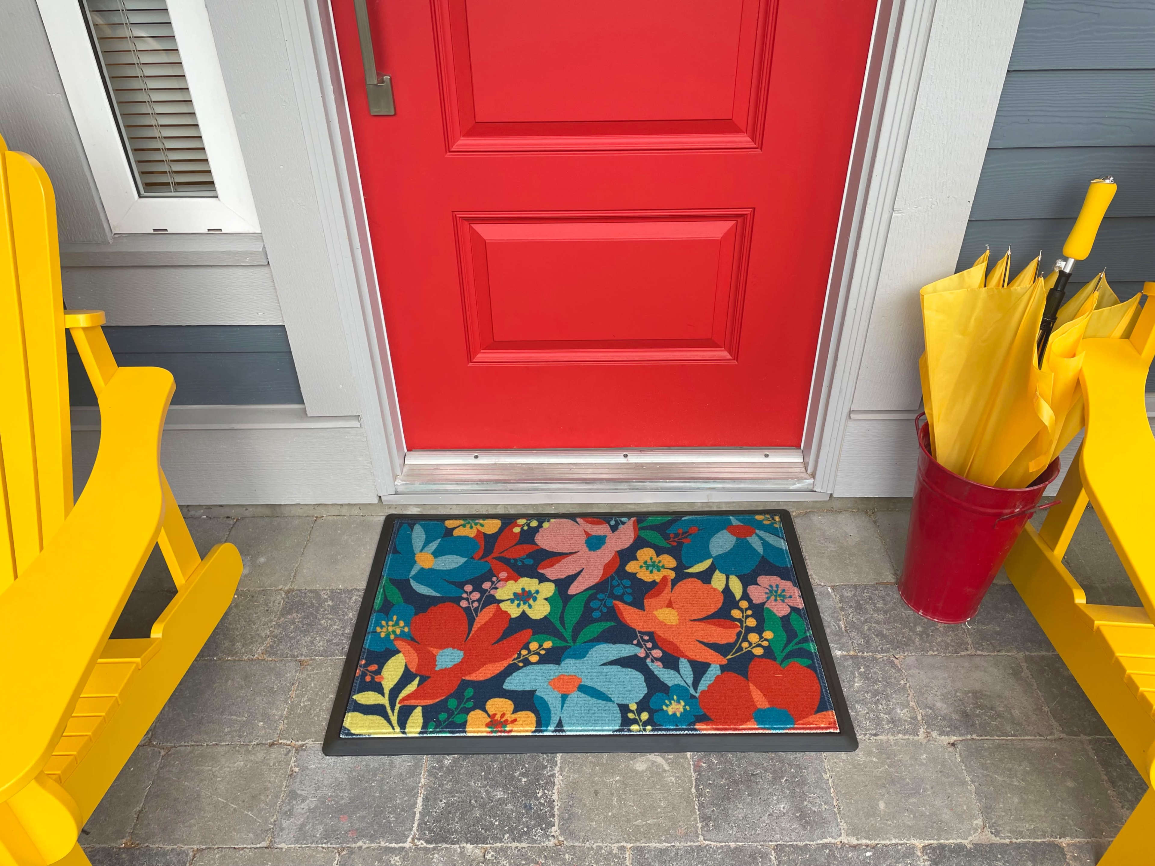 A colorful floral doormat sits in front of a bright red door, flanked by two yellow adirondack chairs and a bucket of yellow umbrellas.