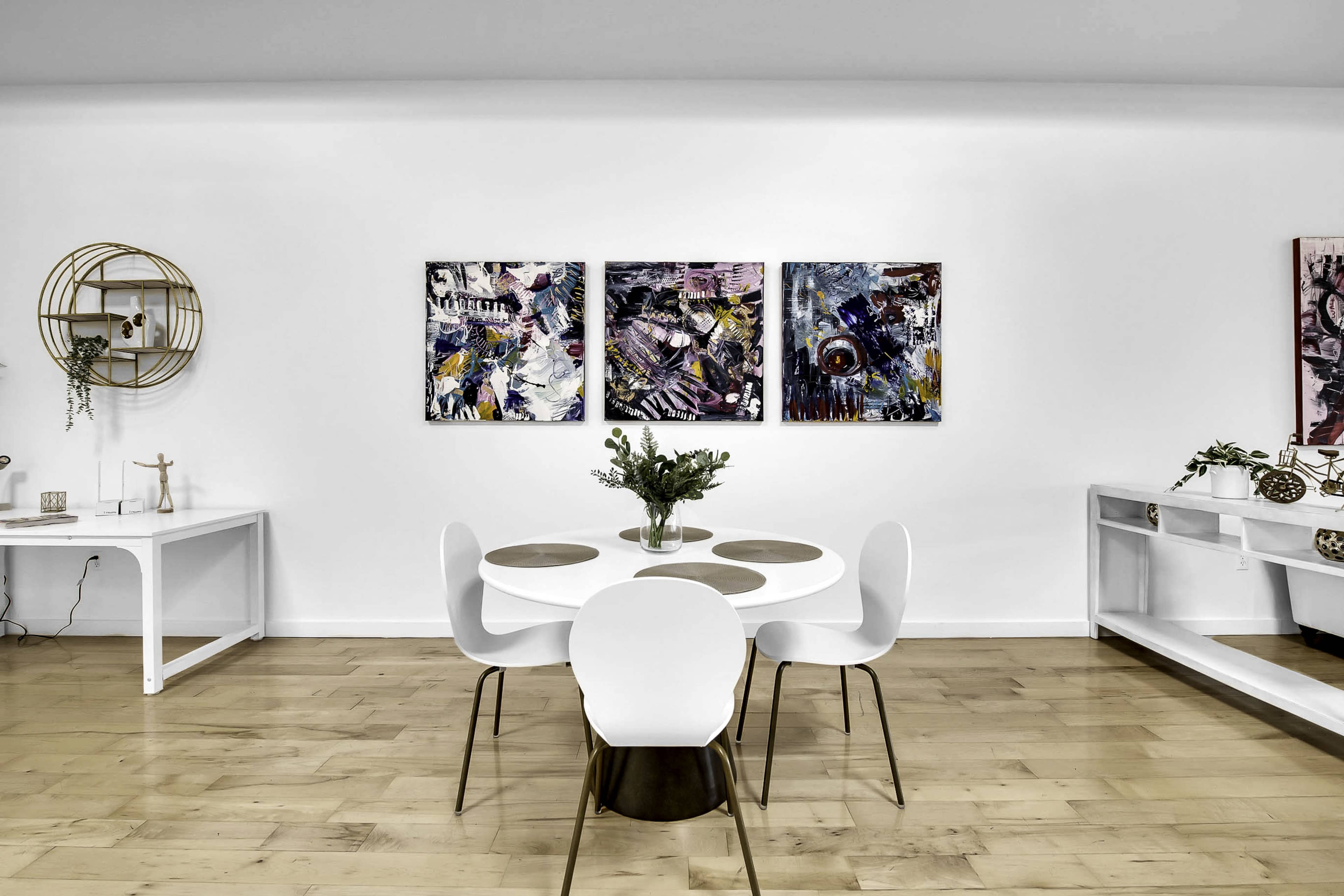 A modern dining area features a round table surrounded by white chairs, with three colorful paintings on the wall and minimalist decor.